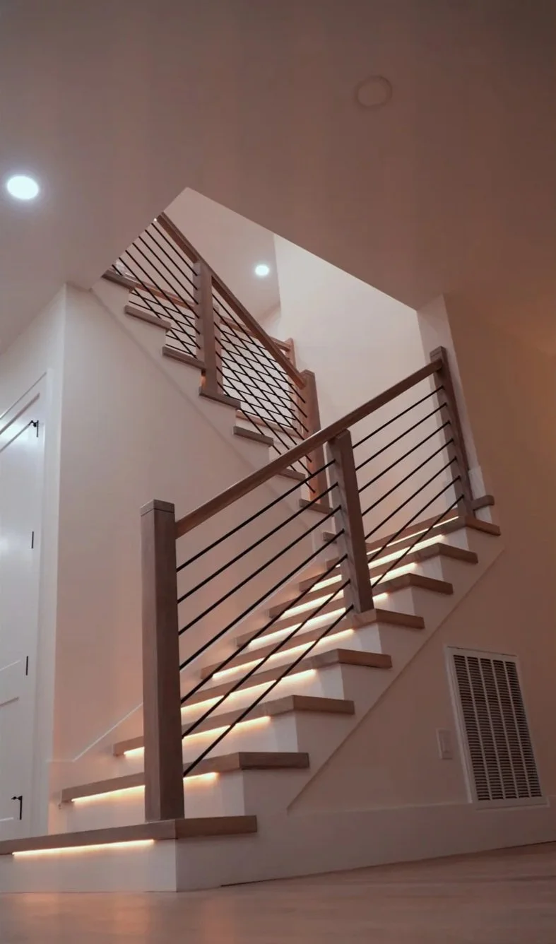 Interior view of a staircase with modern wooden handrails and metal balusters, illuminated by built-in lighting along the steps.
