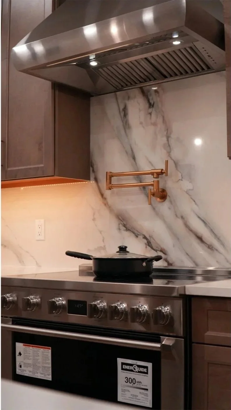 Modern kitchen with marble backsplash, stainless steel range hood, stove with control knobs, black skillet with glass lid on stove, copper towel holder on marble wall, and wooden cabinets.