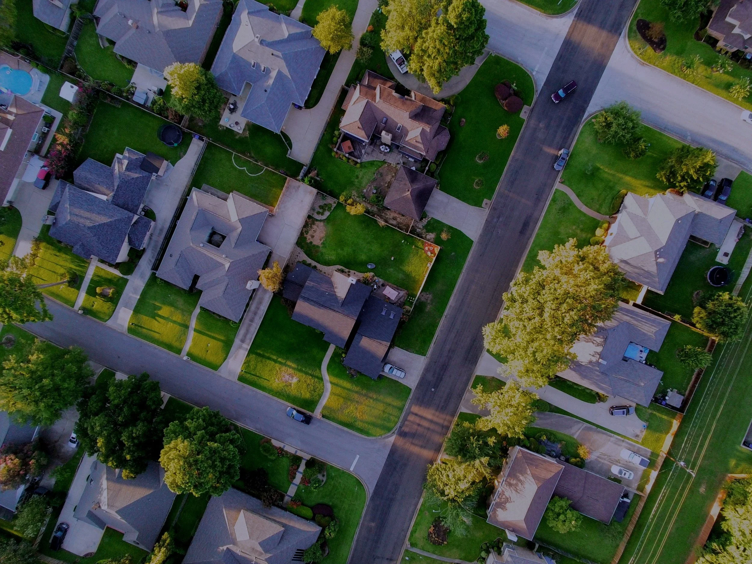 An aerial view of a suburban neighborhood showing houses with gabled roofs, green lawns, trees, driveways, and parked cars along streets.