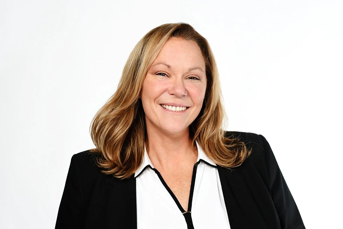 Smiling woman with shoulder-length blonde hair wearing a black blazer and white top, against a white background.