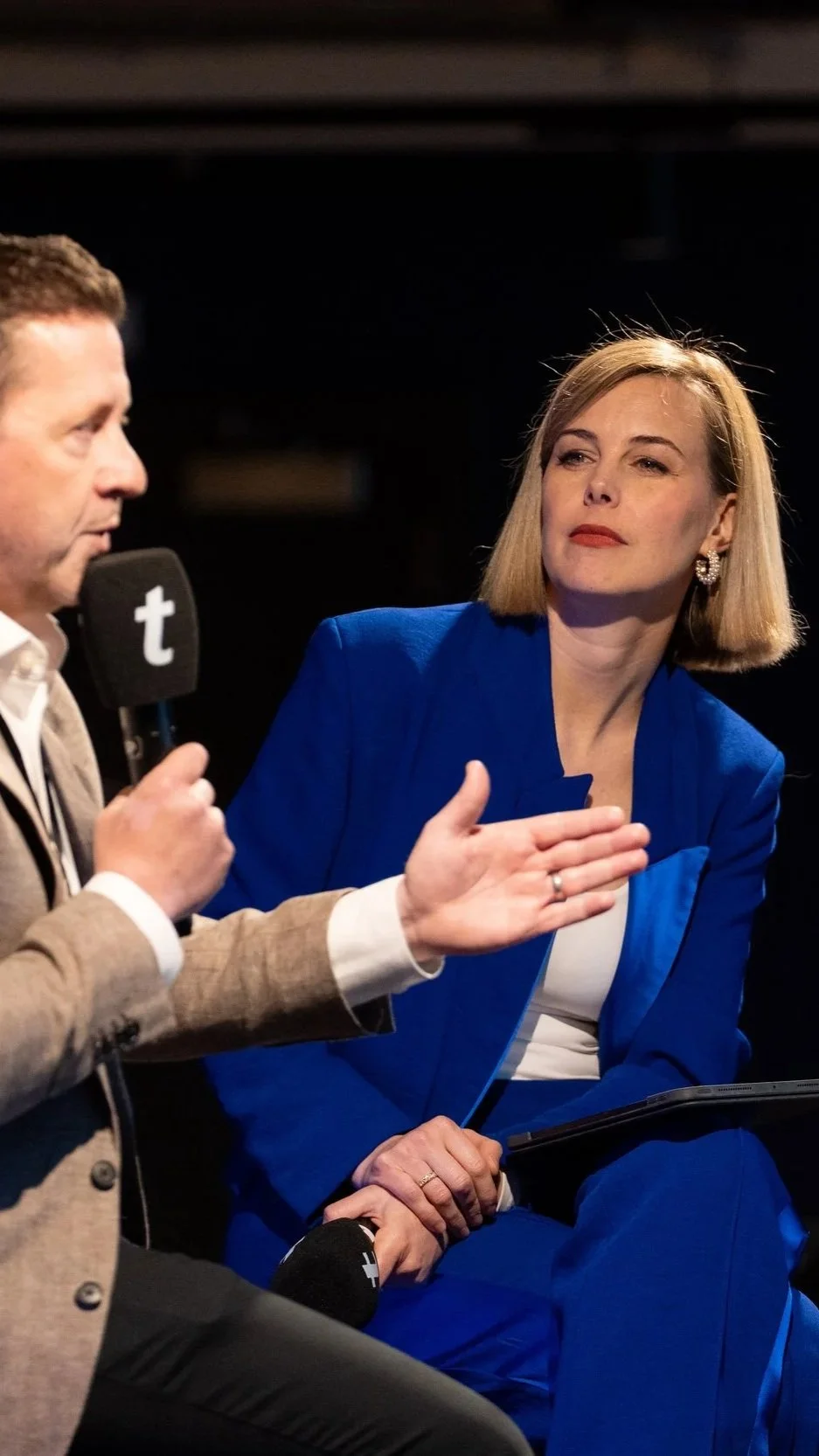 A man holding a microphone with a 't' logo speaking to a woman with blonde hair wearing a blue suit, sitting and listening attentively.