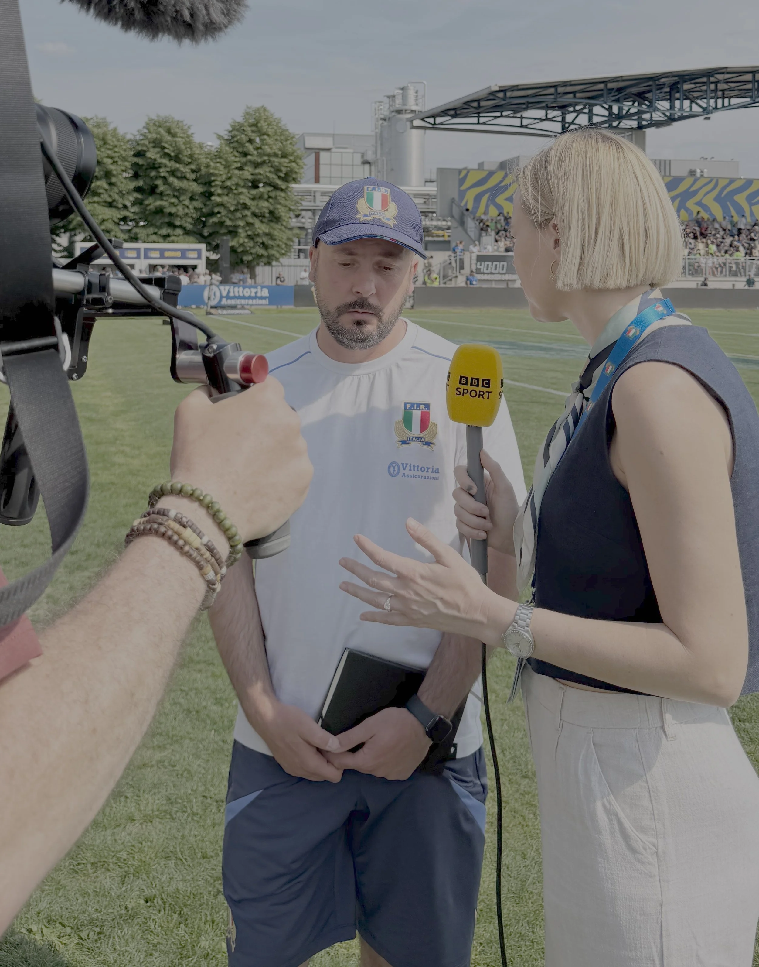 A man being interviewed by a woman at an outdoor event on a sports field. The man is wearing a blue cap and a white T-shirt with Italian flags and sponsors. The woman is holding a yellow BBC Sport microphone and wearing cream pants and a navy sleeveless top with a medal ribbon around her neck. There are trees, a covered grandstand, and an audience in the background.