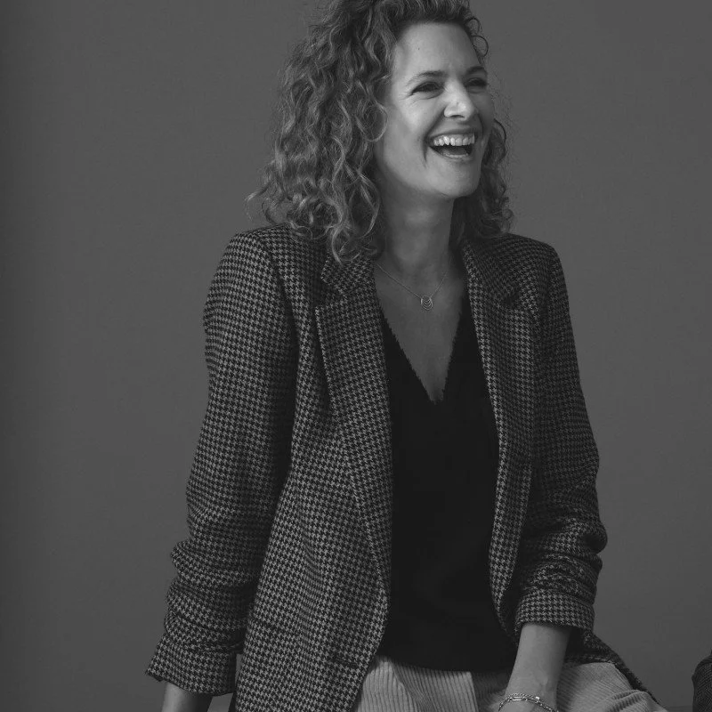 Black and white photo of a woman with curly hair smiling, wearing a patterned blazer over a dark top, against a plain background.