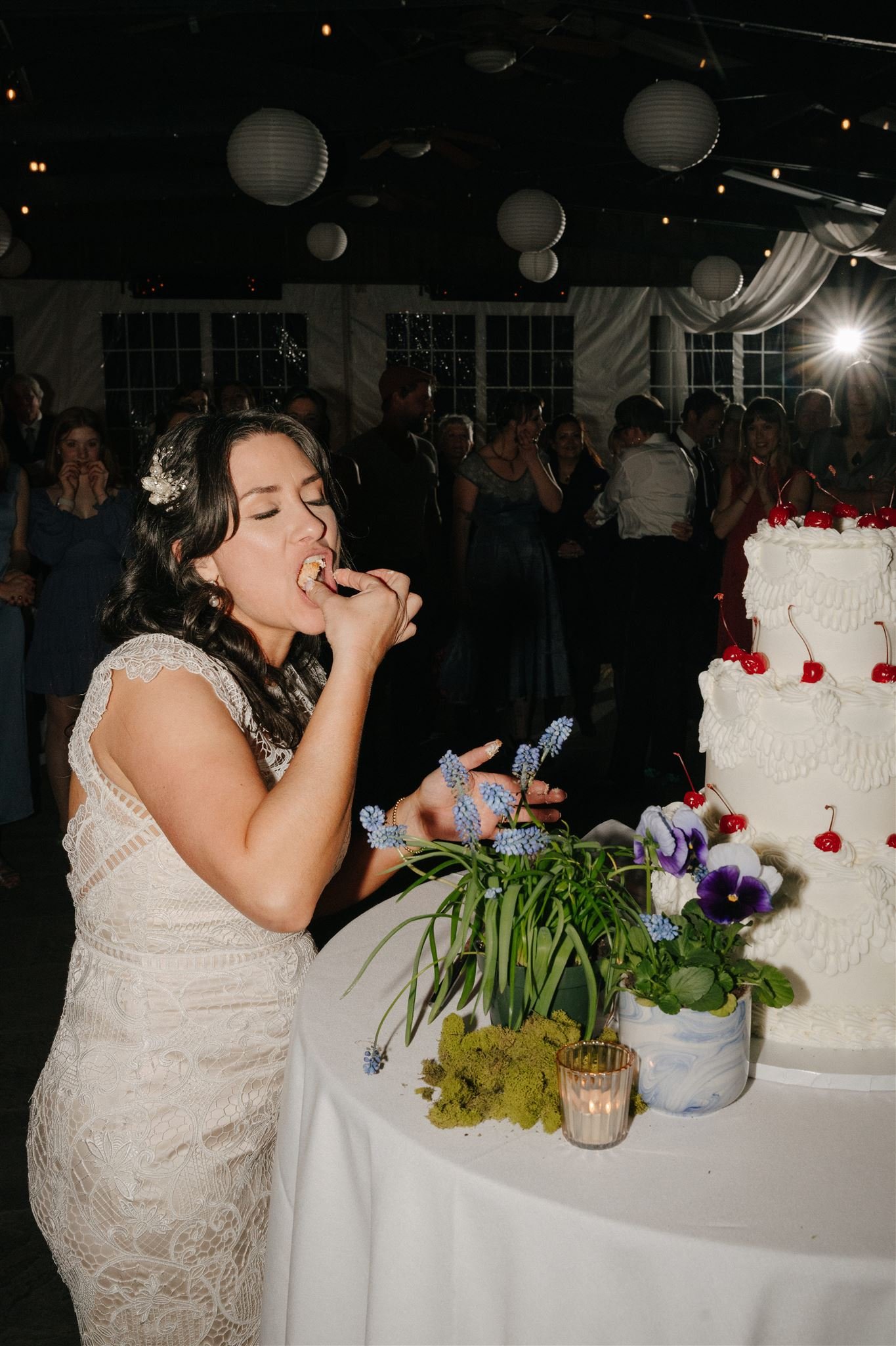 A woman in a white lace dress at a wedding reception, standing beside a wedding cake decorated with cherries and white icing, with a bouquet of purple and white flowers and a lit candle in a glass holder on the table in front of her. Other guests are visible in the background, clapping and celebrating inside a decorated venue with white drapes, paper lanterns, and string lights.