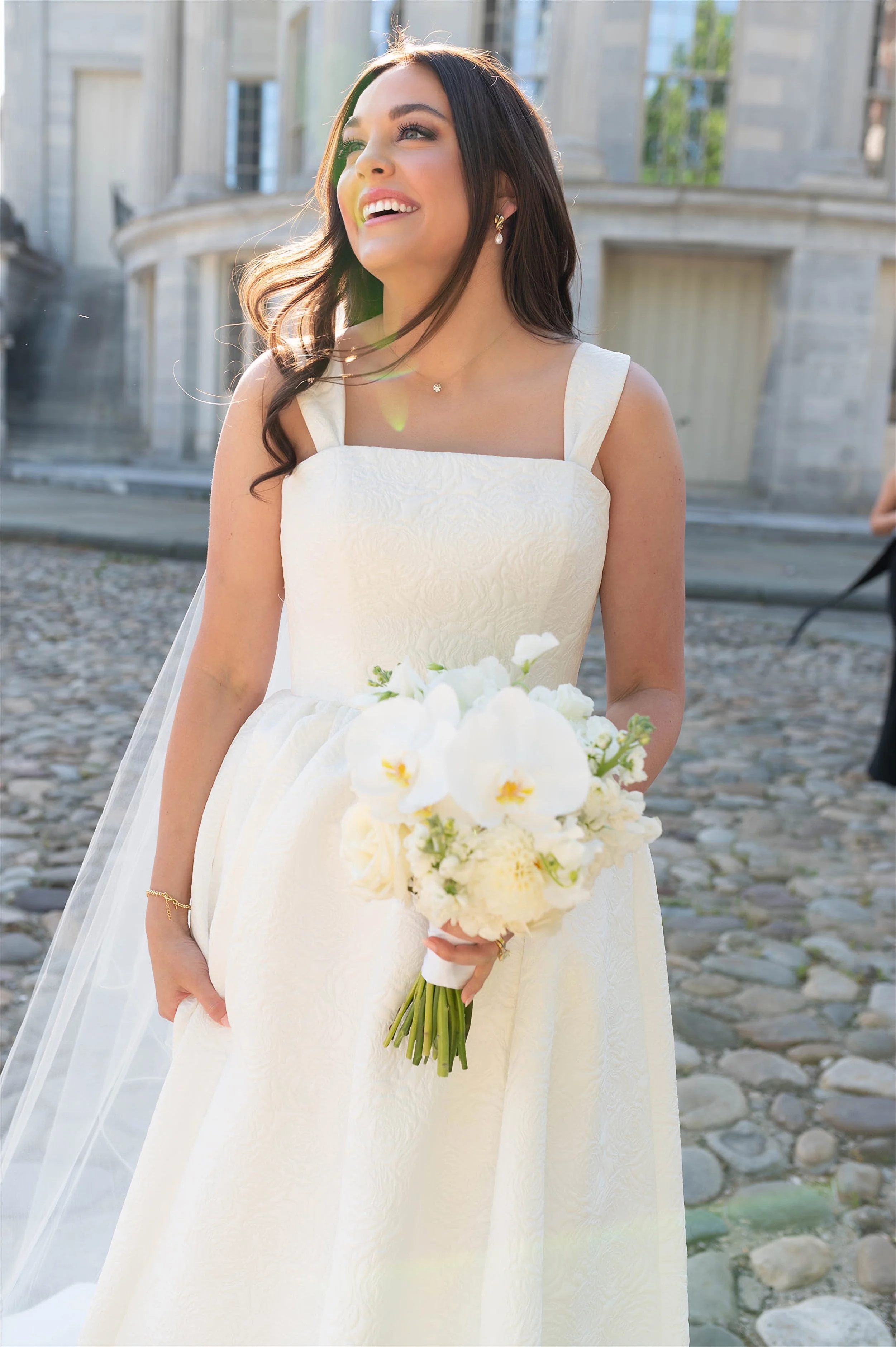 A woman in a white wedding dress holding a bouquet of white and yellow flowers, smiling outdoors in front of a historic building with stone architecture.