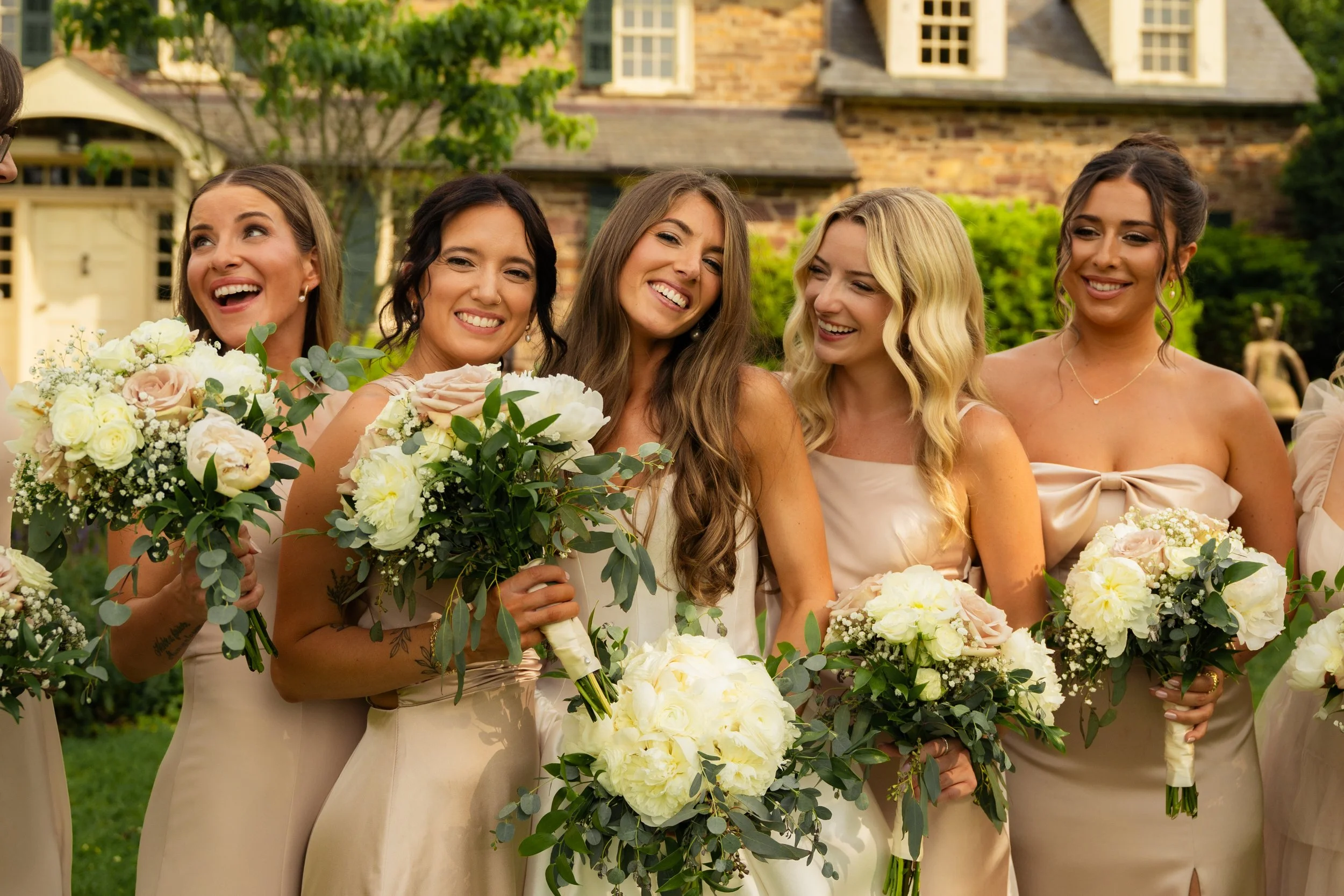 A group of bridesmaids holding bouquets of white and pink flowers, standing outdoors in front of a brick house, smiling and enjoying a wedding celebration.