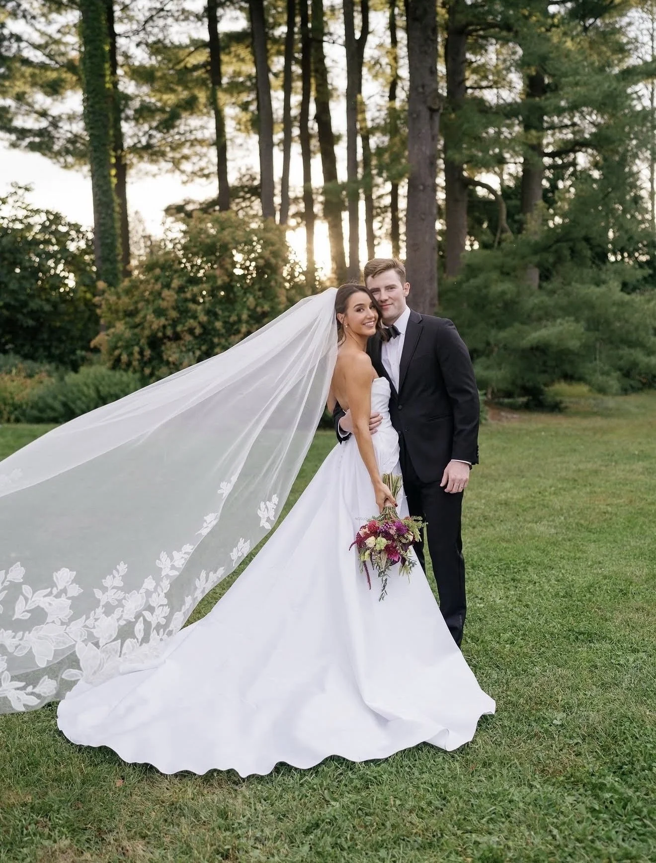 A bride and groom in wedding attire standing outdoors on grass with trees in the background at sunset. The bride wears a white gown with a long lace veil, and the groom wears a black tuxedo. The bride holds a bouquet of flowers, and they are smiling.