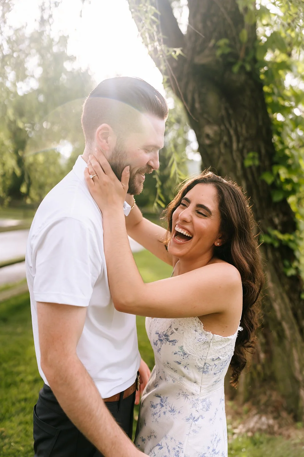 A happy couple sharing a joyful moment outdoors near a large tree, with sunlight filtering through the leaves, both smiling and embracing each other.