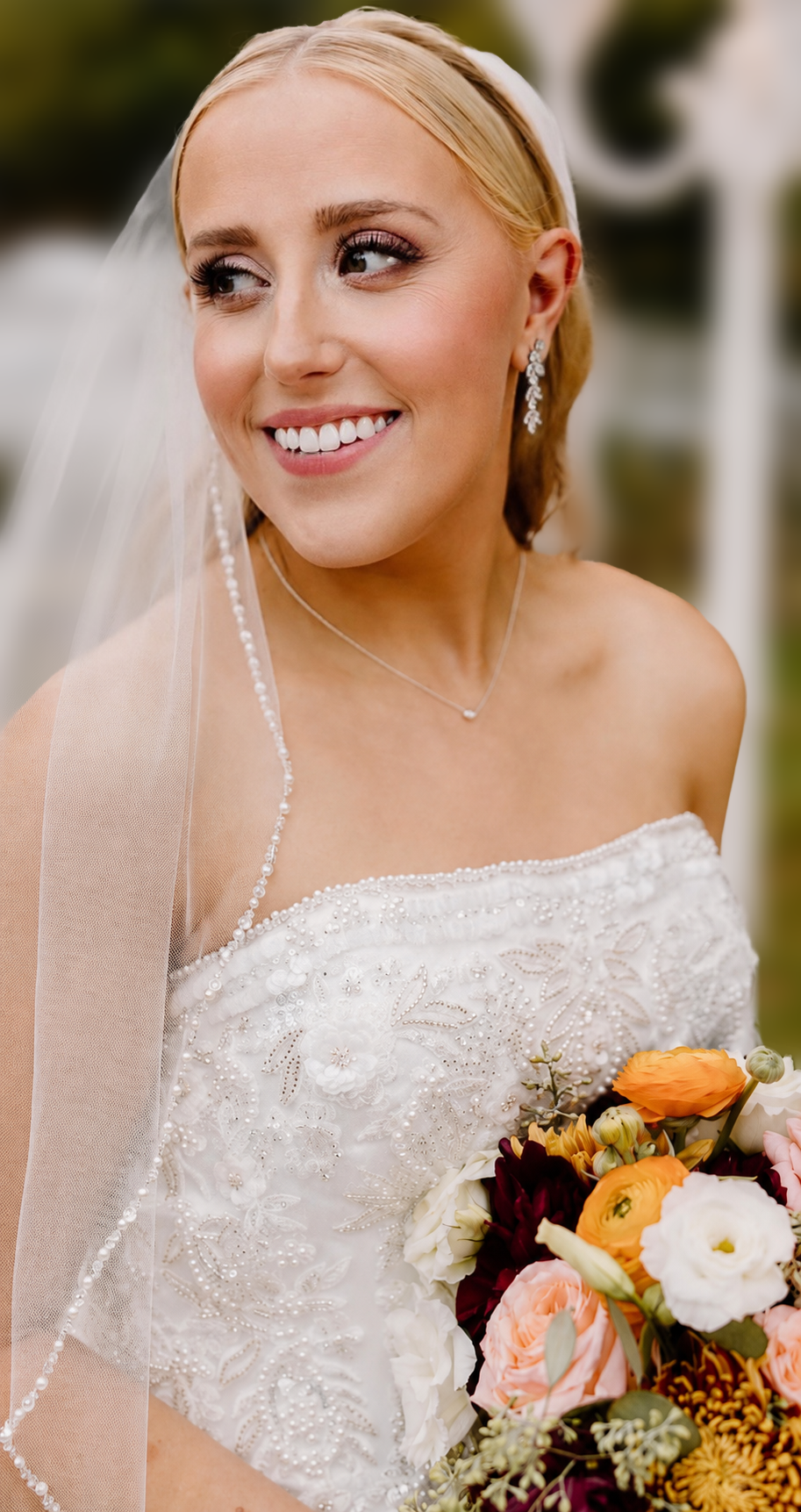 A smiling bride wearing a wedding dress and veil, holding a bouquet of colorful flowers, with earrings, necklace, and styled blonde hair.
