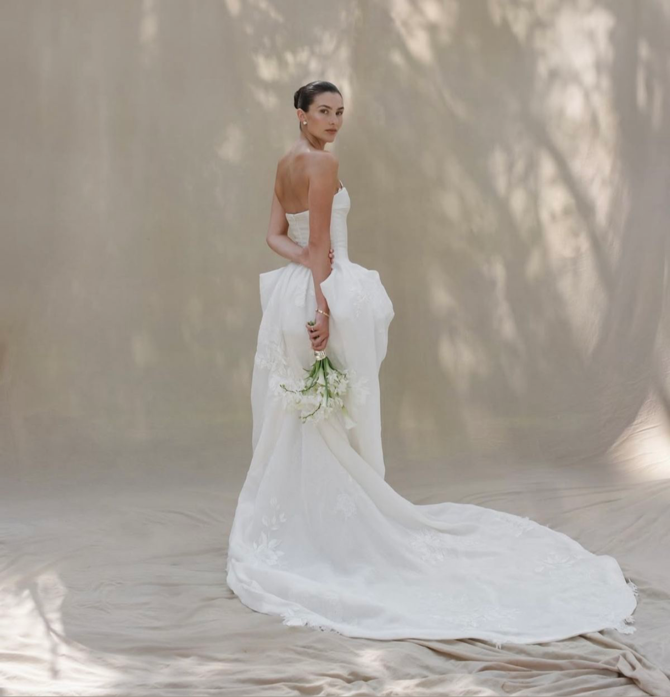 A woman in a white wedding dress holding a small bouquet of white flowers, standing against a neutral backdrop.
