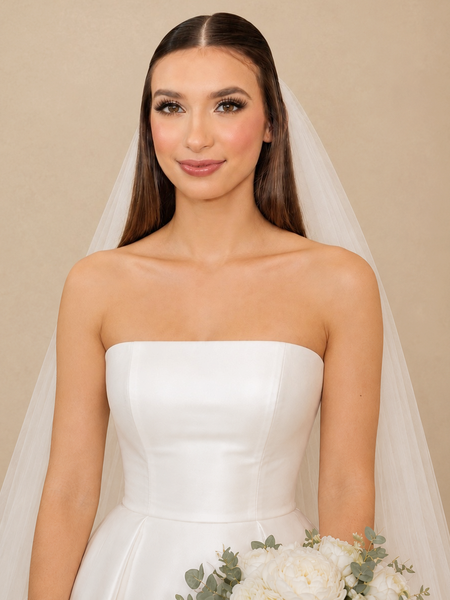 Brunette bride in strapless white wedding dress holding a bouquet of white flowers with eucalyptus, smiling softly, with a beige background.