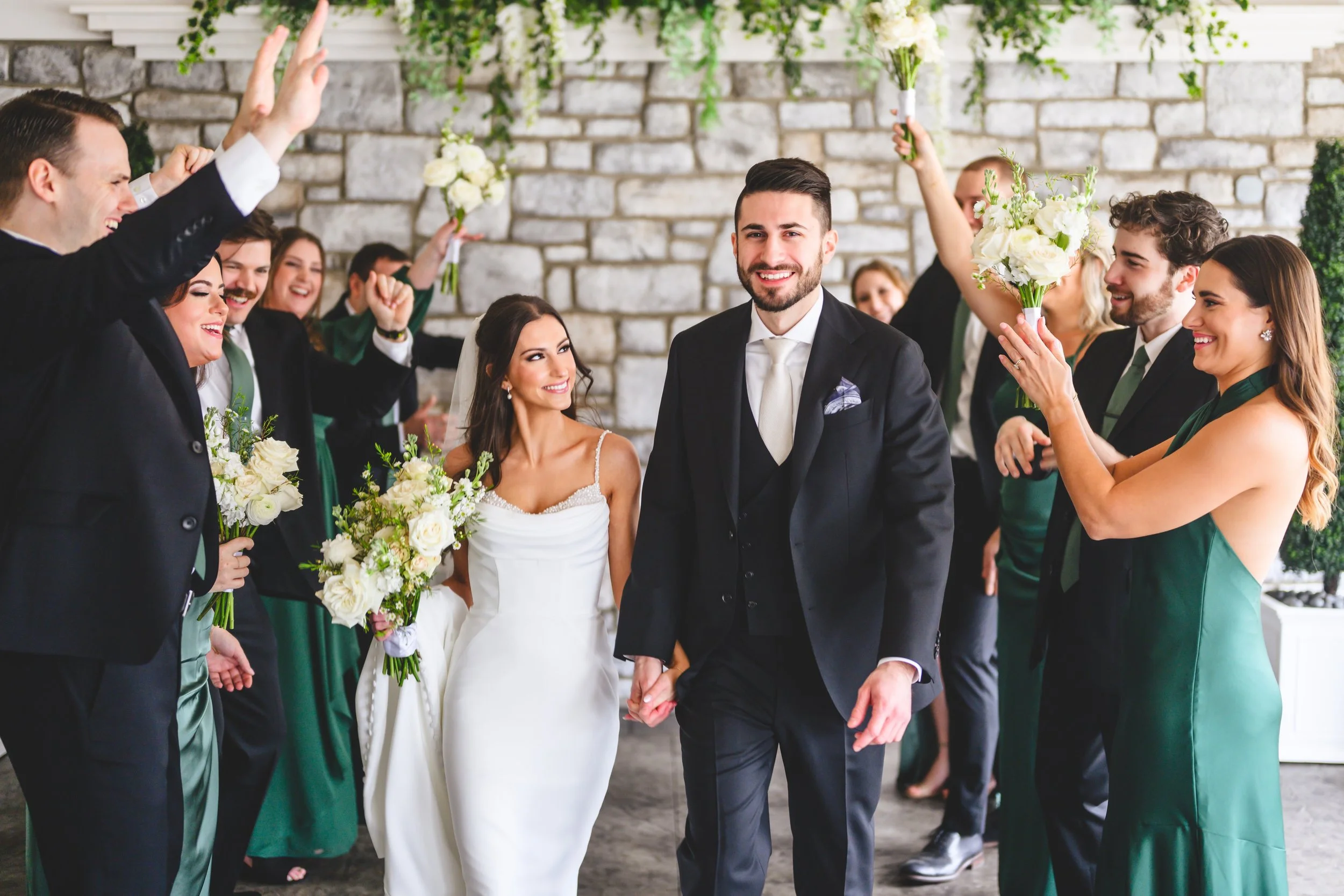 A wedding party celebrating indoors. The bride and groom hold hands in the center, surrounded by bridesmaids and groomsmen holding bouquets and dressed in formal attire. The background features a stone wall and greenery.