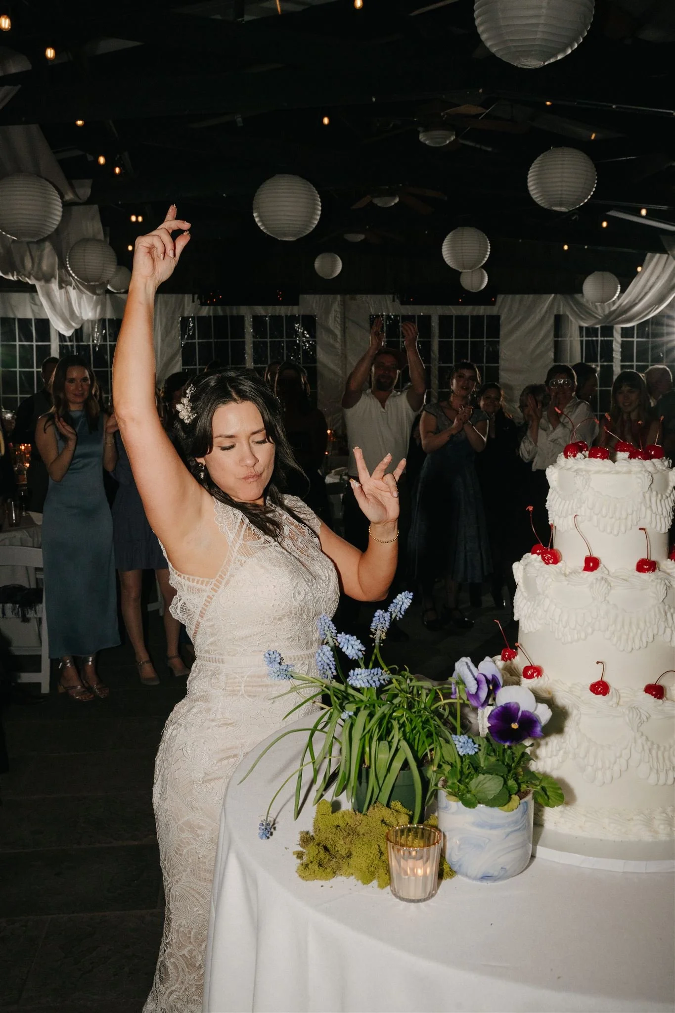 A woman in a white lace dress at a wedding reception, standing beside a wedding cake decorated with cherries and white icing, with a bouquet of purple and white flowers and a lit candle in a glass holder on the table in front of her. Other guests are visible in the background, clapping and celebrating inside a decorated venue with white drapes, paper lanterns, and string lights.