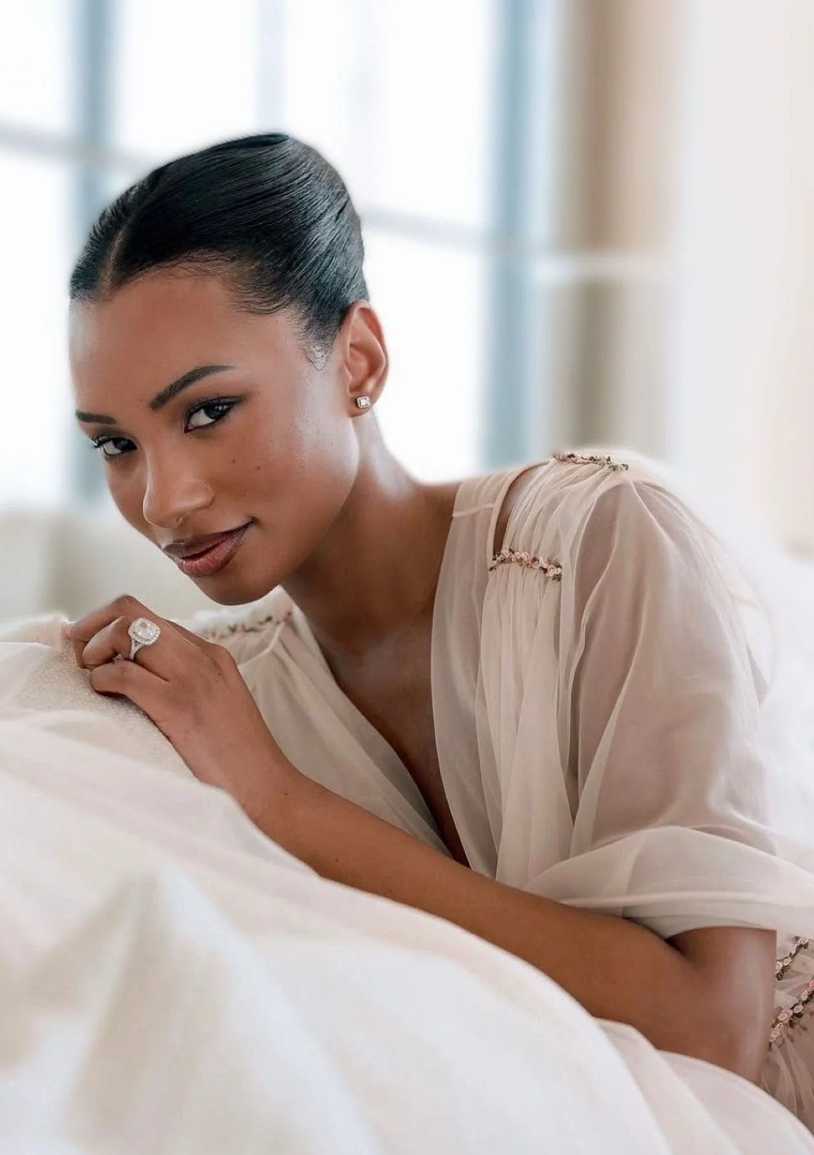 A woman with dark hair pulled back, wearing a light sheer blouse with embroidery details, lying on a bed with a soft, white blanket, looking at the camera with a gentle smile.