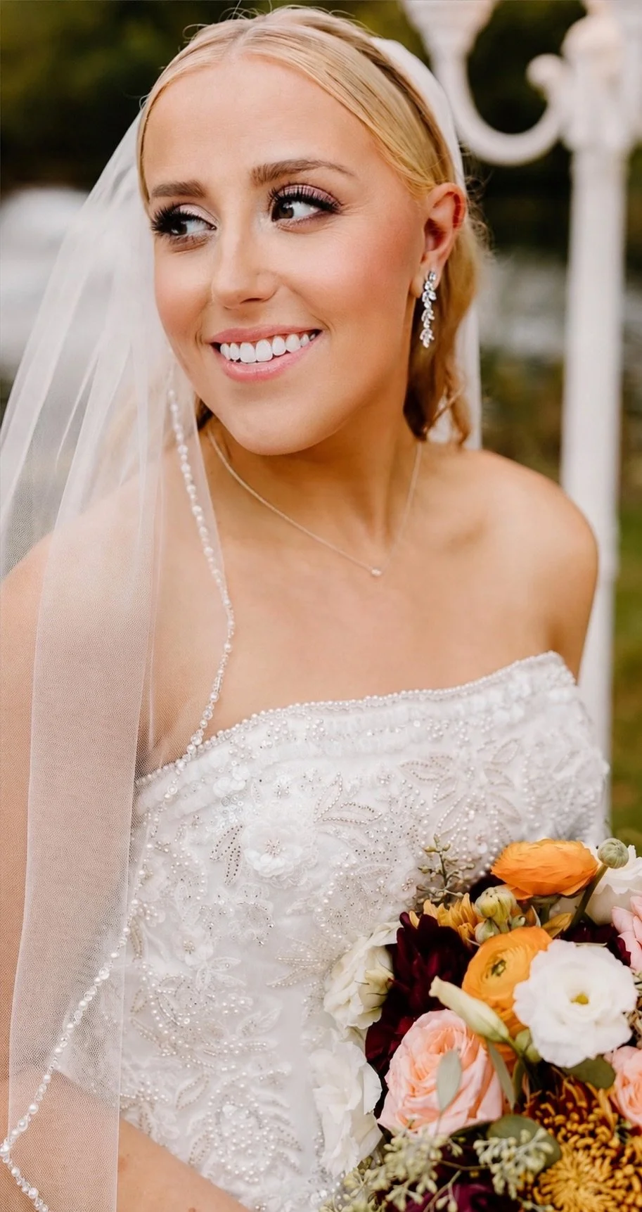 Close-up of a smiling bride in a white wedding dress with intricate embroidery, holding a bouquet of orange, white, and dark red flowers, with a veil and jewelry, outdoors with greenery and white garden decor in the background.