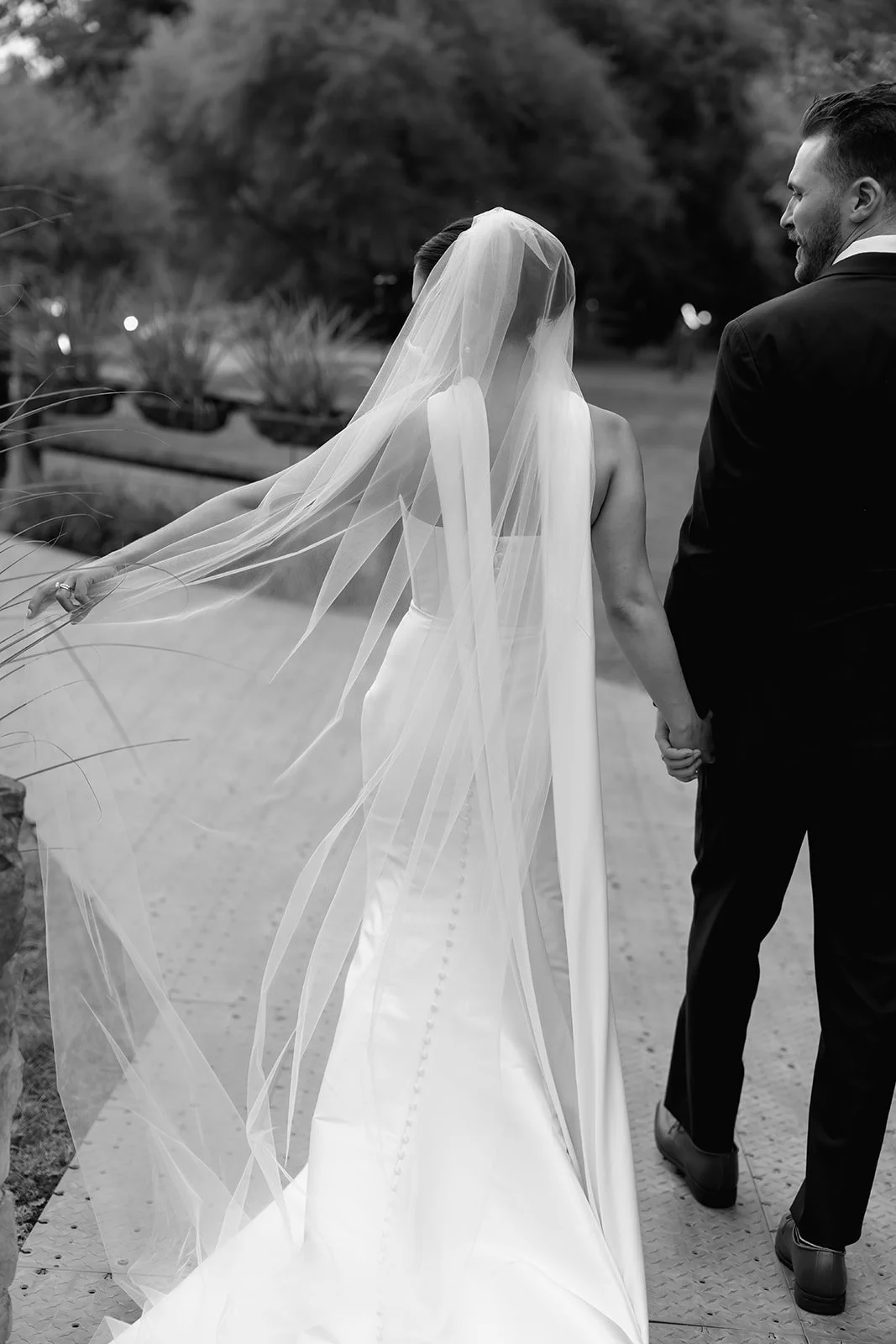 A bride and groom walk hand in hand outdoors, holding hands, with the bride wearing a wedding dress and veil, and the groom in a suit, with trees and landscaping in the background.
