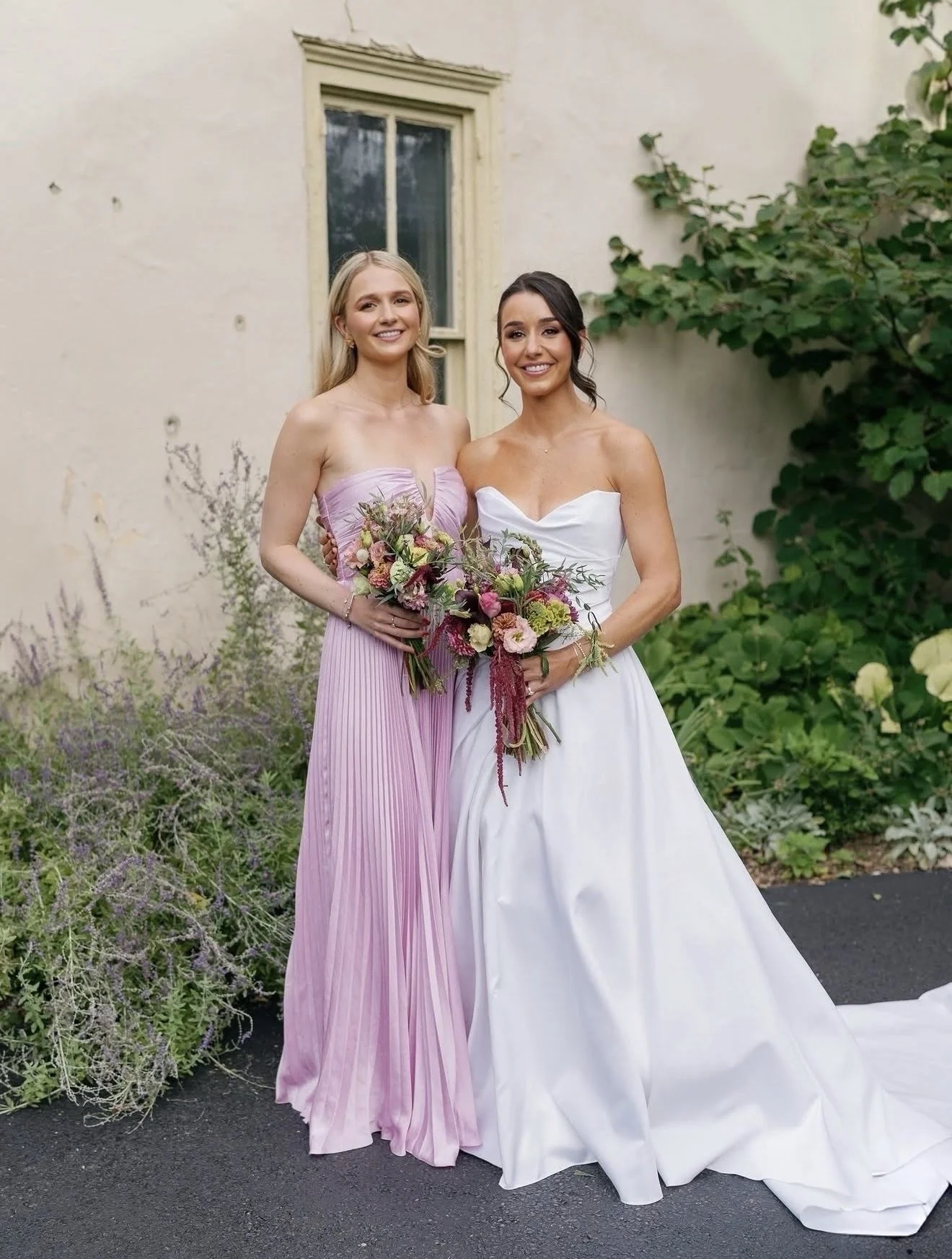 Two women in wedding attire standing outside in a garden, holding bouquets of flowers, smiling at the camera.