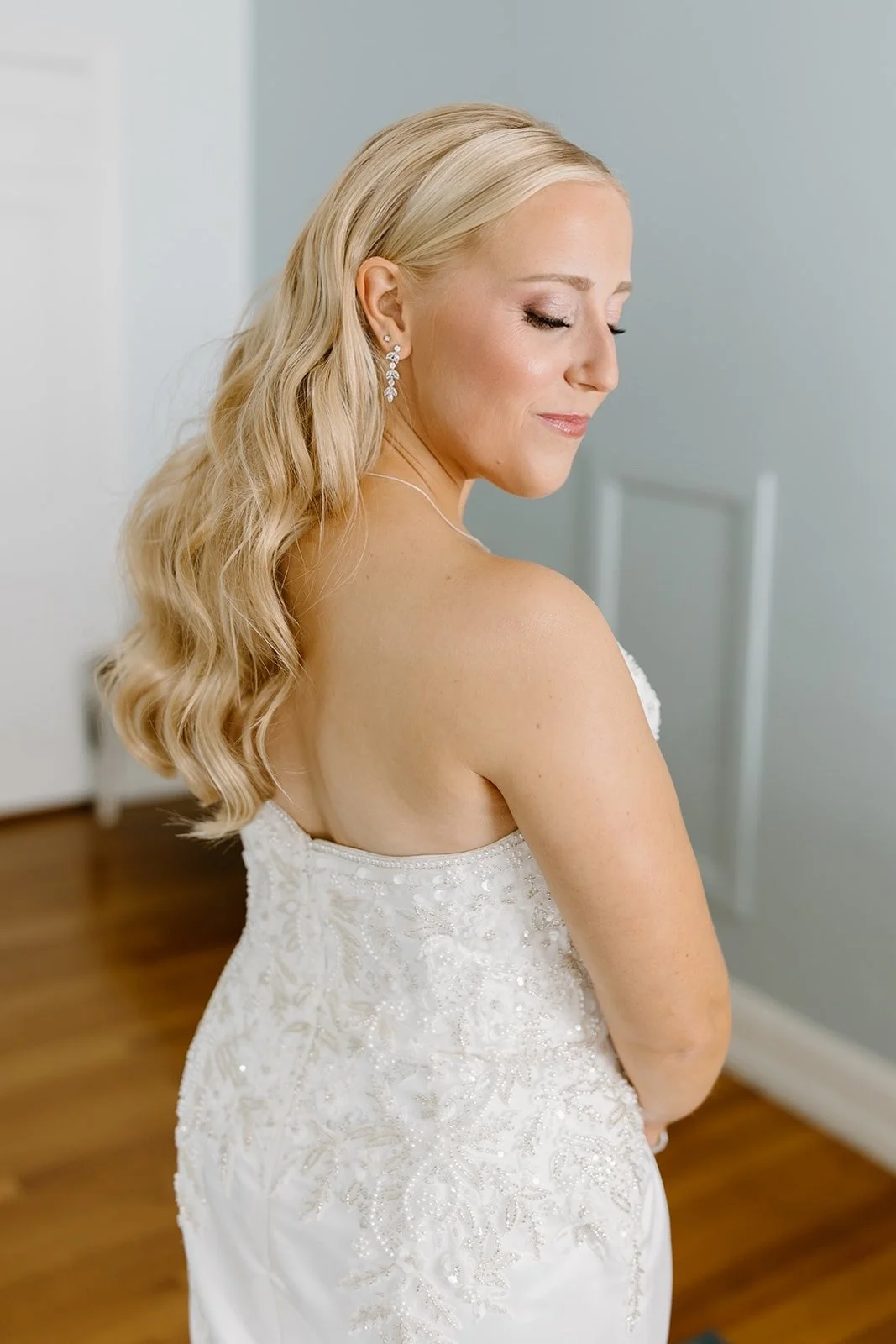 A woman with long blonde wavy hair and earrings in a white wedding dress, smiling with eyes closed.