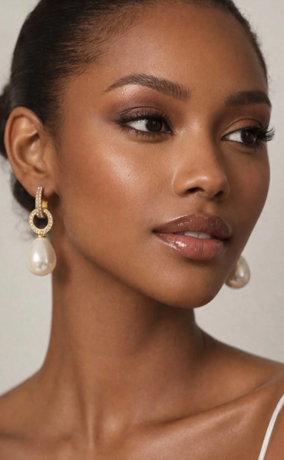 Close-up of a woman with smooth brown skin and elegant makeup, wearing gold earrings with pearl drops.