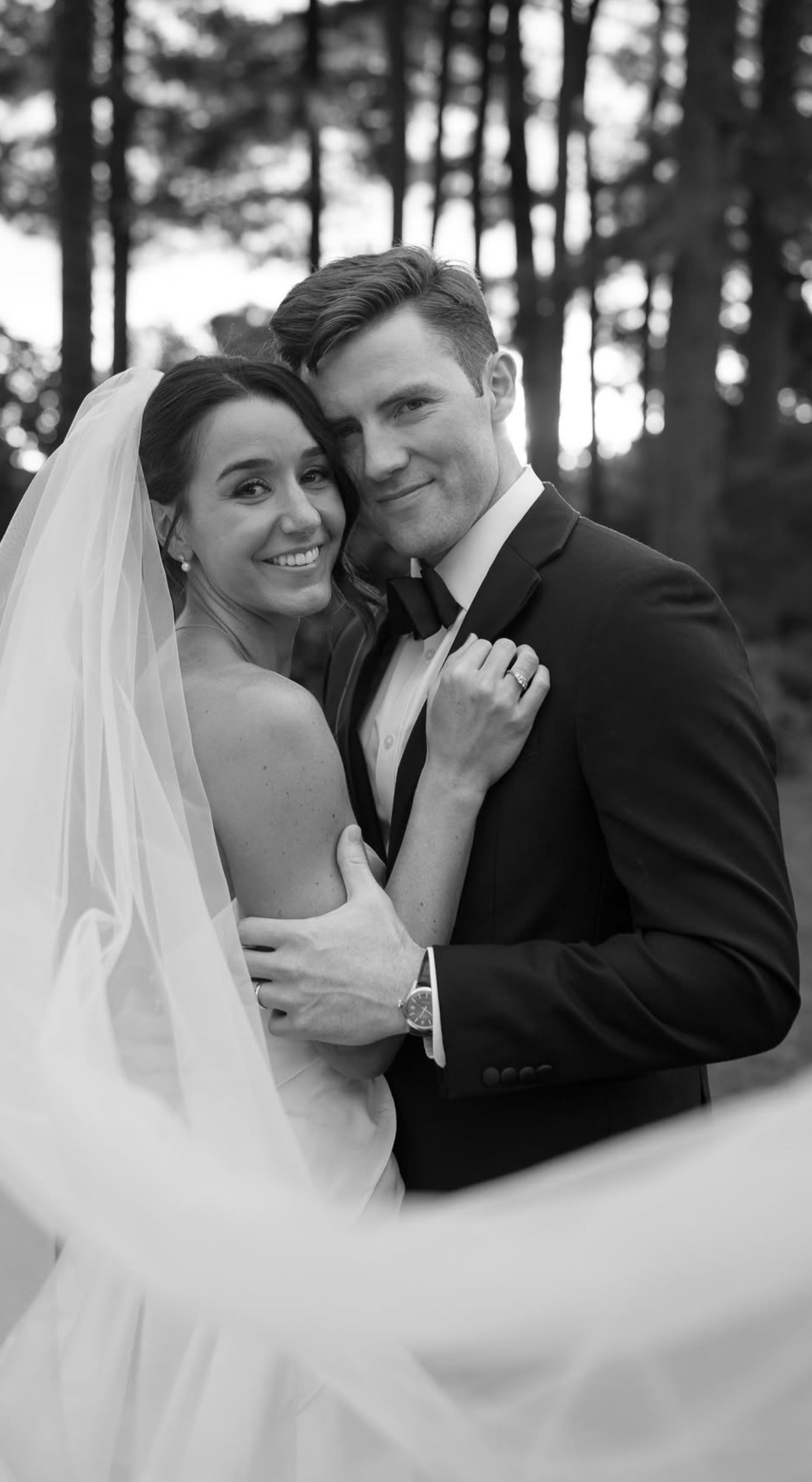 A bride and groom dressed in wedding attire, standing close together outdoors, smiling and embracing each other. The photo is black and white with a background of trees.
