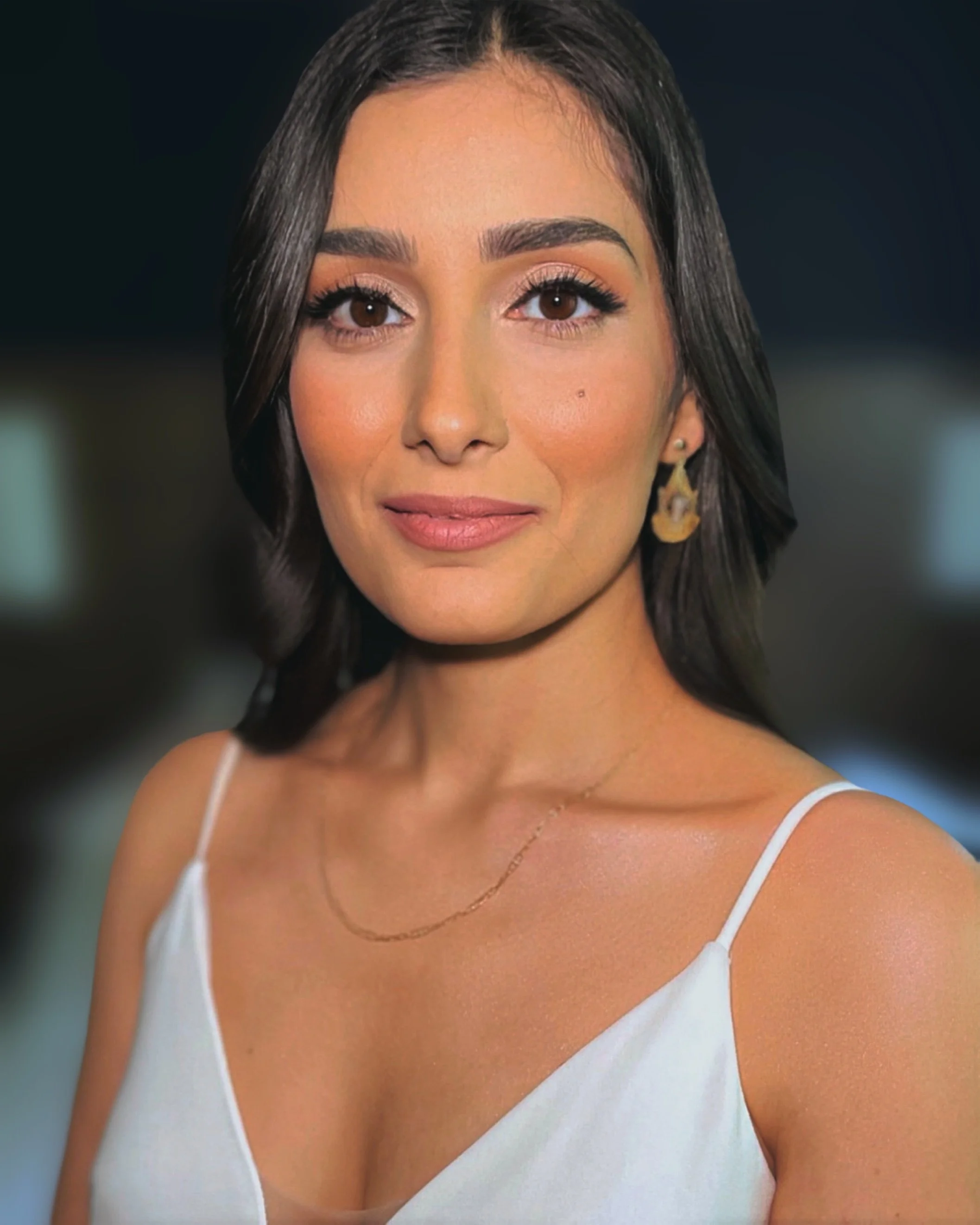 Close-up of a woman with dark brown hair styled over her shoulders, wearing a white spaghetti strap top, with makeup including neutral eyeshadow, mascara, and pink lipstick, and gold earrings.