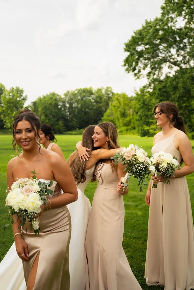 A group of women in beige dresses celebrating outdoors, holding bouquets of white and pink flowers, with green trees and grass in the background.