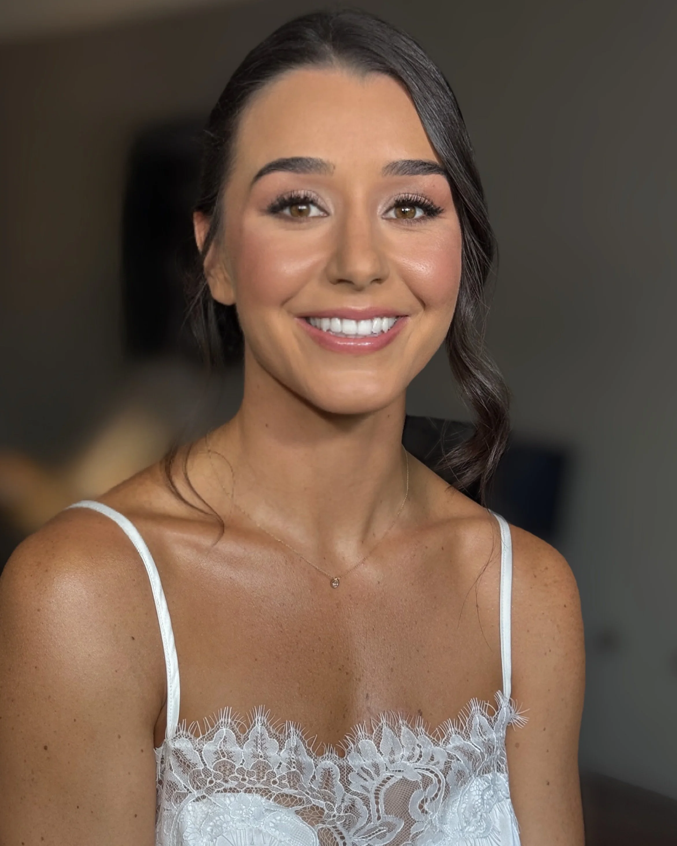 Close-up of a smiling woman with brown hair styled in loose waves, wearing a white lace dress with thin straps and a delicate necklace, standing in a softly lit indoor setting.