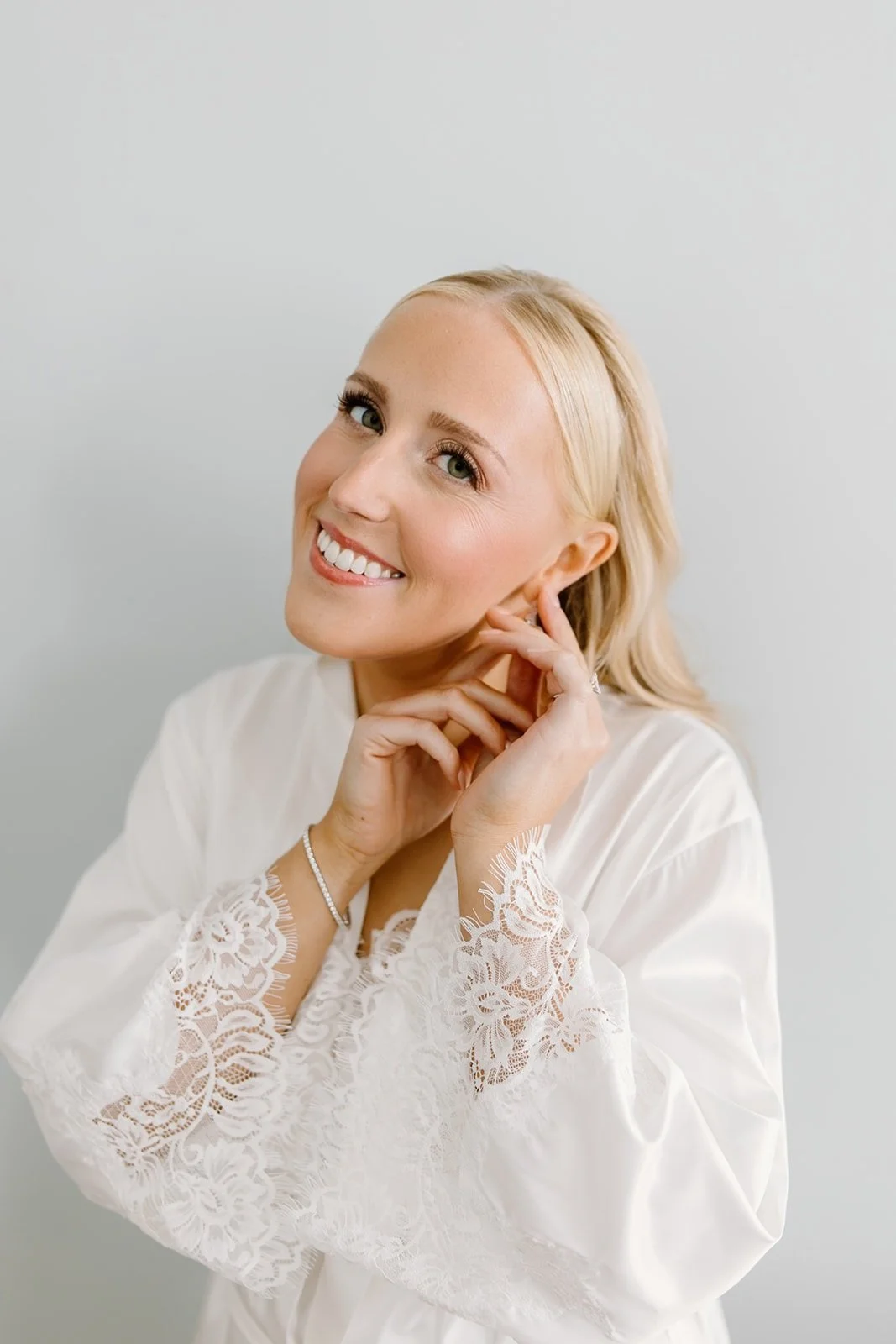 A smiling blonde woman with blue eyes wearing a white lace robe, touching her face and earrings, standing against a plain light background.