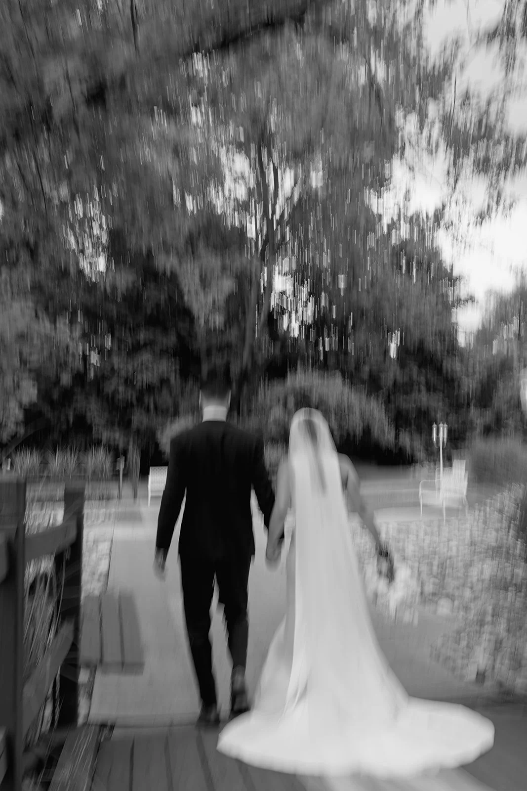 Black and white photo of a bride and groom walking hand in hand on a wooden bridge outdoors, with trees in the background, during evening or dusk.