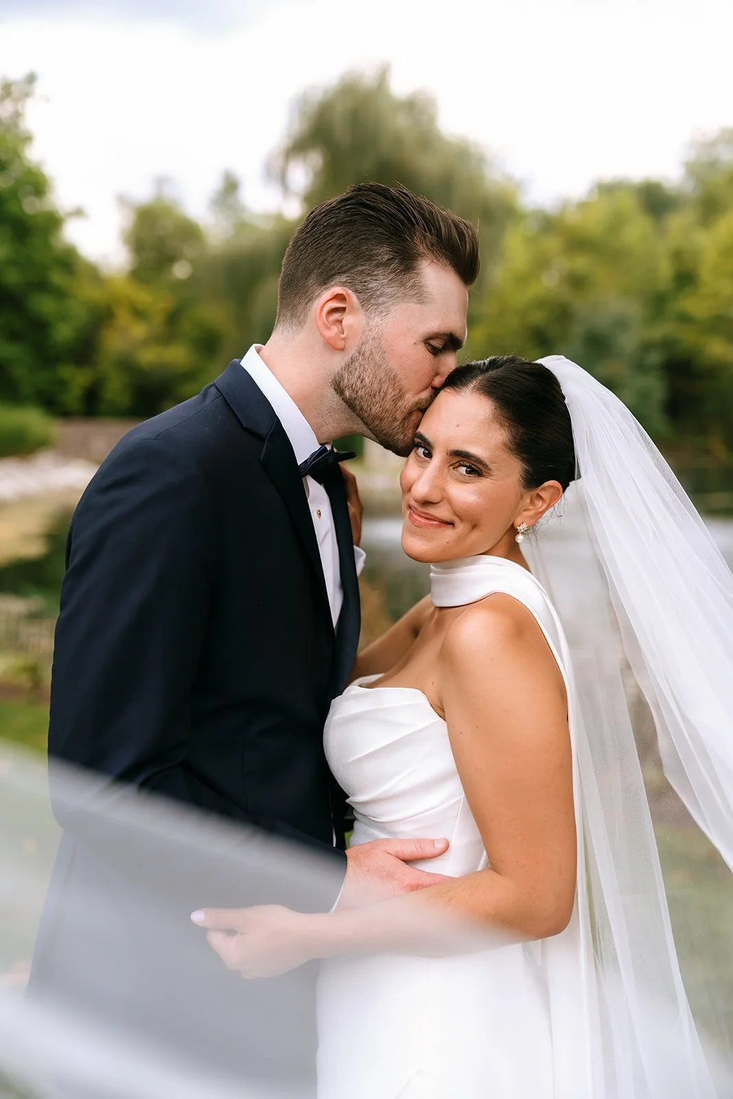A newlywed couple outdoors, with the groom kissing the bride's forehead, as she smiles at the camera. The groom is in a black tuxedo, and the bride is in a white wedding dress with a veil, surrounded by trees and greenery.