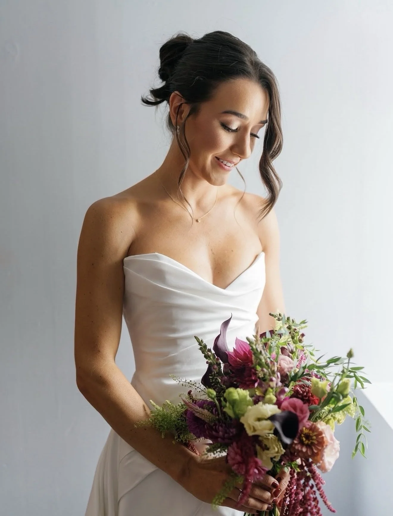 A woman in a strapless white dress holding a large bouquet of colorful flowers, smiling softly with her eyes closed, standing against a light gray background.
