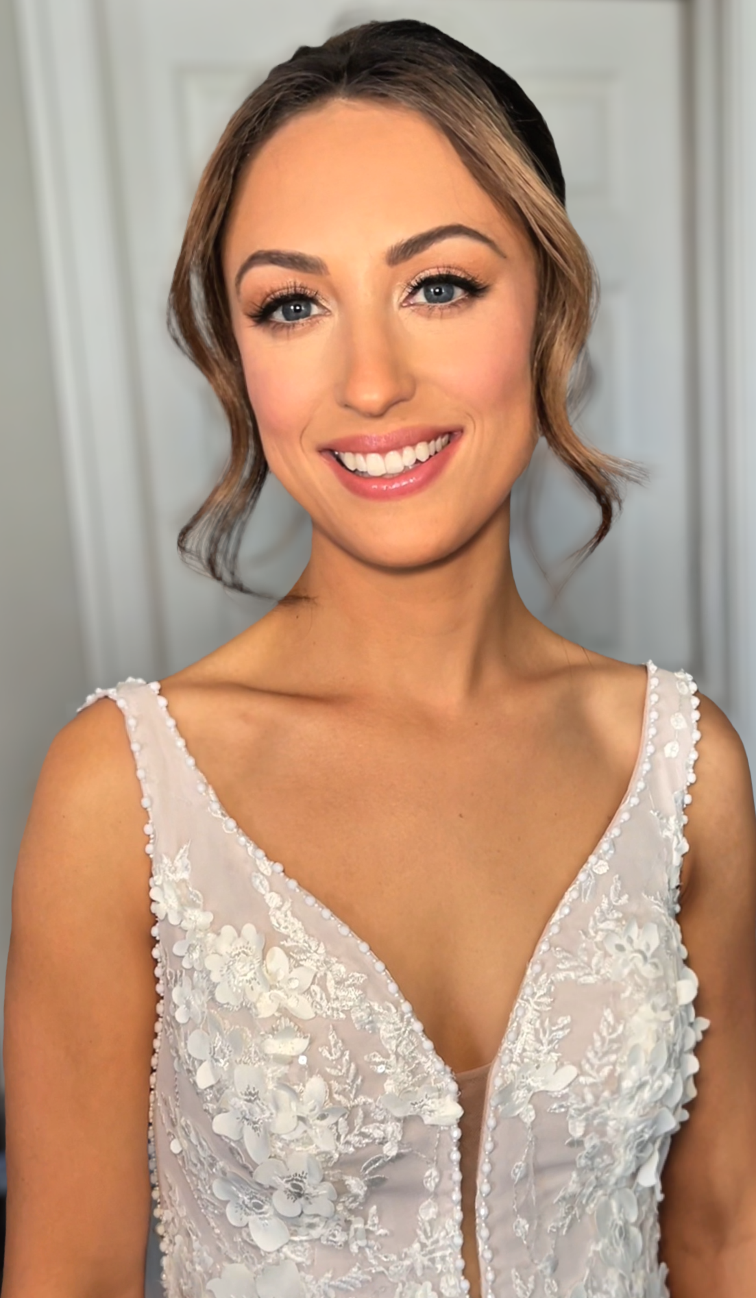 A woman smiling wearing a white dress with floral embroidery, standing in front of a plain background.
