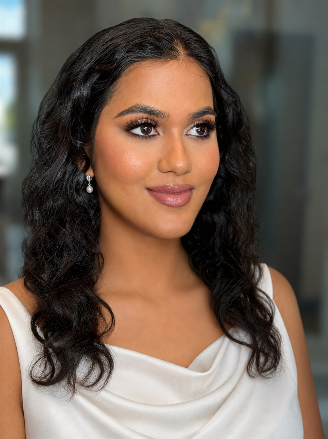 A woman with long curly black hair, wearing pearl earrings and a white sleeveless dress, smiling softly with a blurred modern building background.