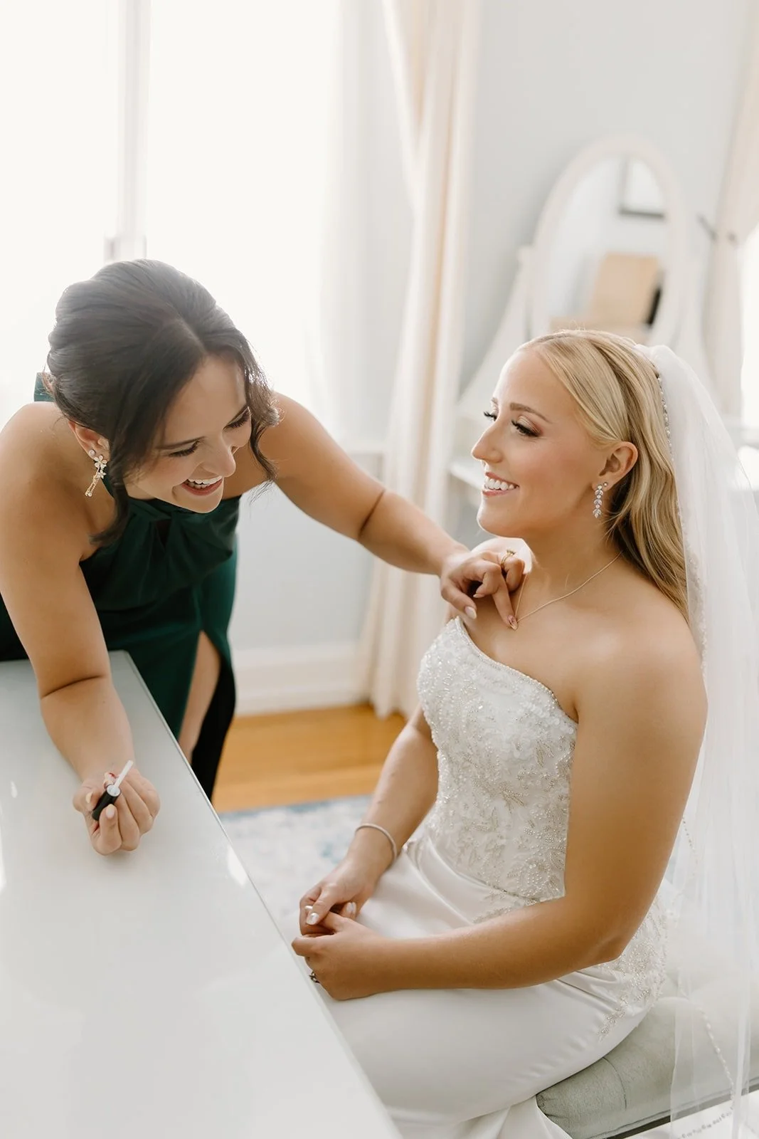 Bridal bride smiling and sitting in white wedding dress while her makeup artist helps her with makeup in a bright room.