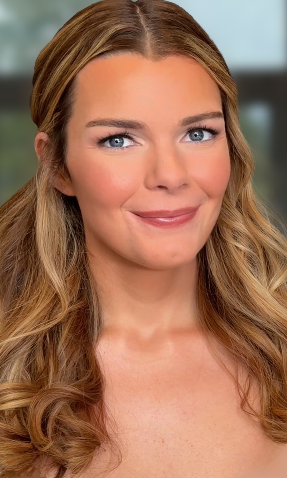 Close-up of a woman with long wavy light brown hair, blue eyes, and a subtle smile, with a blurred background.