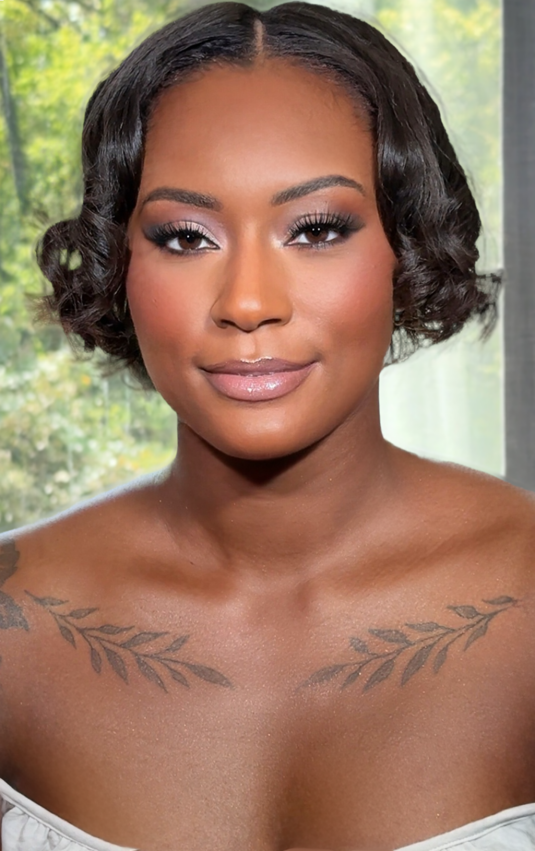 Close-up of a woman with short, wavy black hair, wearing makeup, and tattoos of leaves across her collarbones.