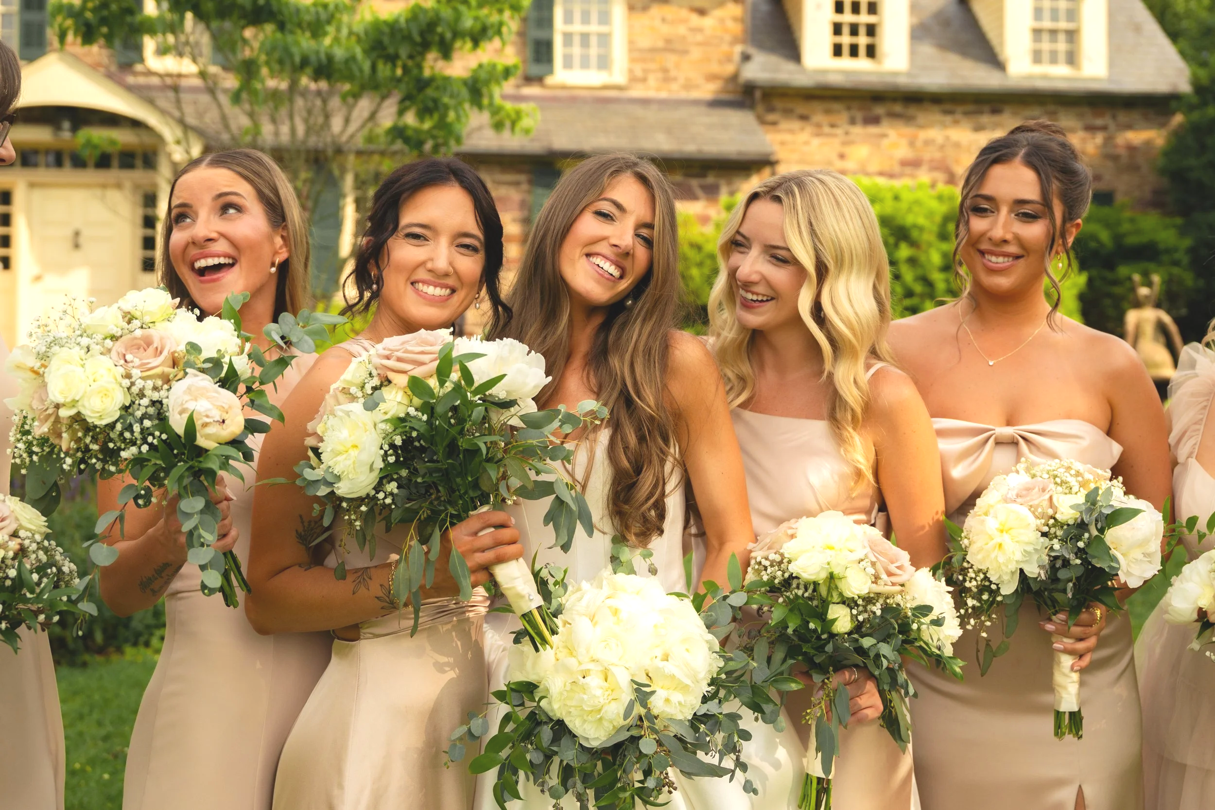 A group of women in elegant dresses holding bouquets of flowers, smiling and celebrating outdoors in front of a house with trees and greenery.
