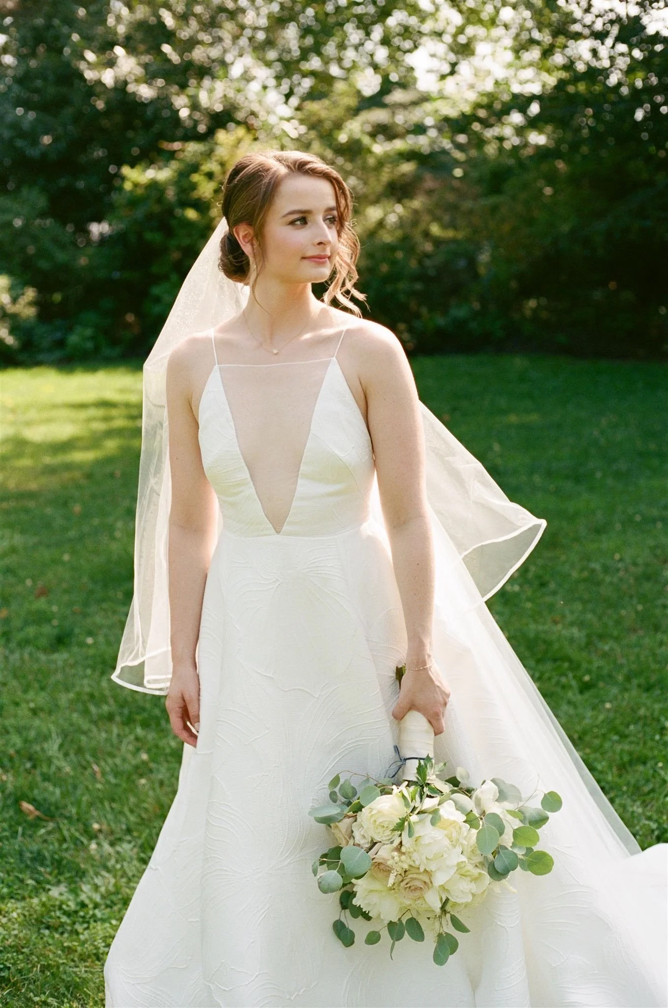 A young woman in a white wedding dress standing outdoors on a grassy area with green trees in the background, holding a bouquet of white flowers and greenery.
