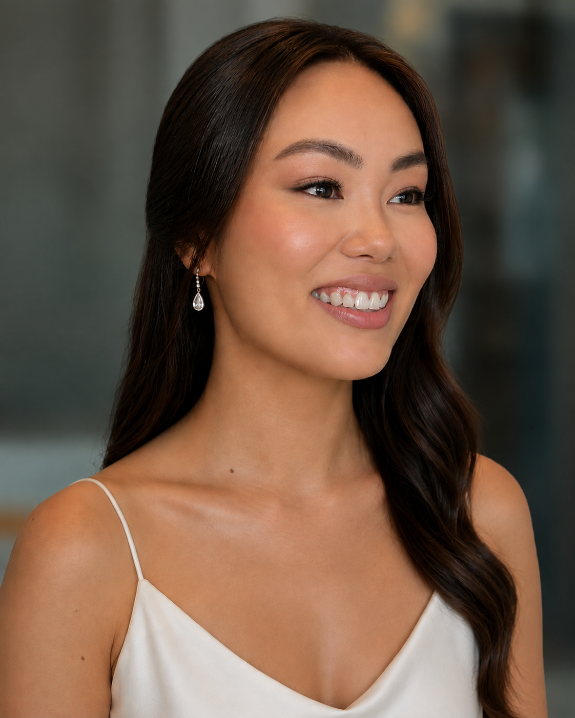 A young woman with long dark hair smiling, wearing pearl earrings and a white dress with thin straps.