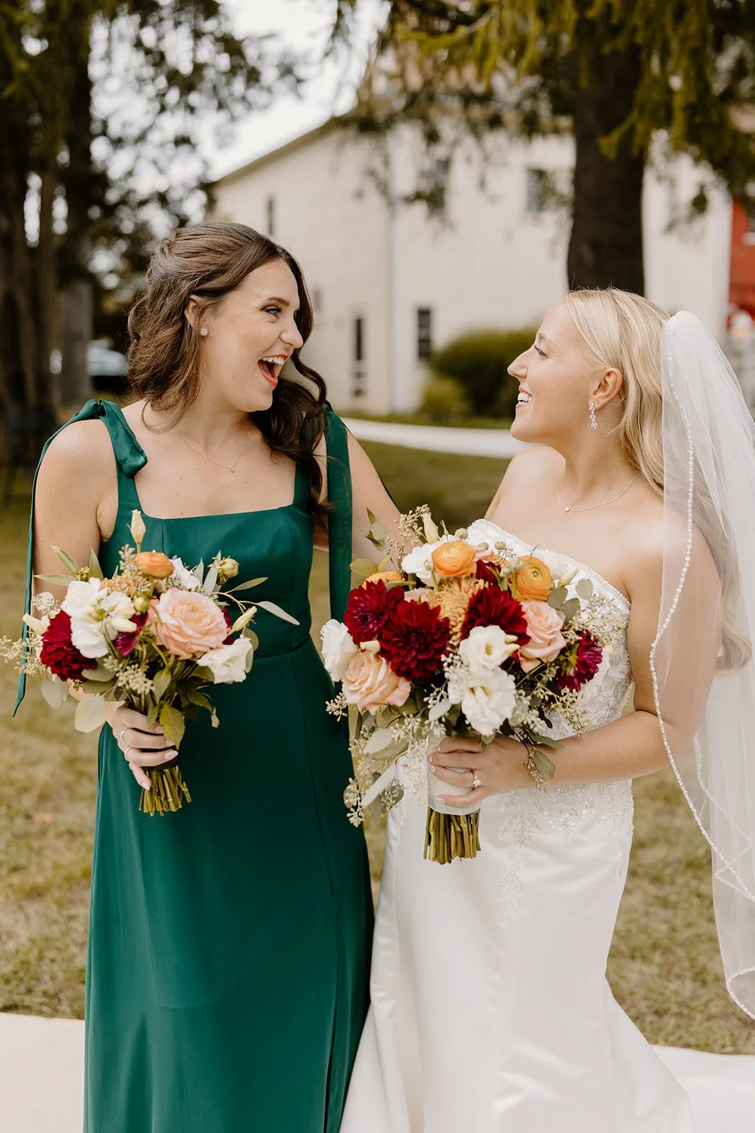 A bride and a bridesmaid sharing a joyful moment outdoors, holding bouquets of flowers. The bride wears a white wedding gown with a veil, and the bridesmaid wears a green dress. They are smiling and looking at each other warmly.