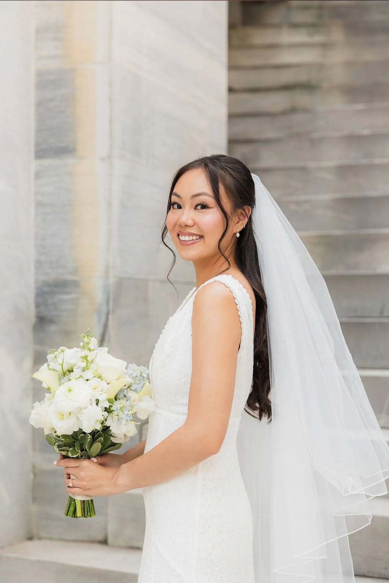 A smiling bride with long dark hair, wearing a white wedding dress and veil, holding a bouquet of white flowers, standing indoors near a staircase and a stone wall.