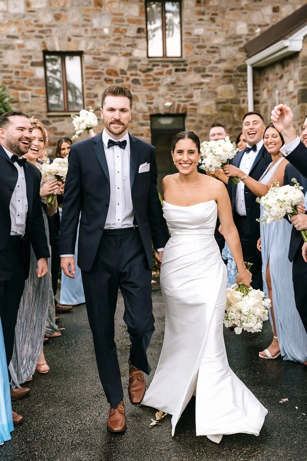 A newlywed couple walking down the aisle, surrounded by friends and family who are celebrating and holding bouquets, with a brick wall background.