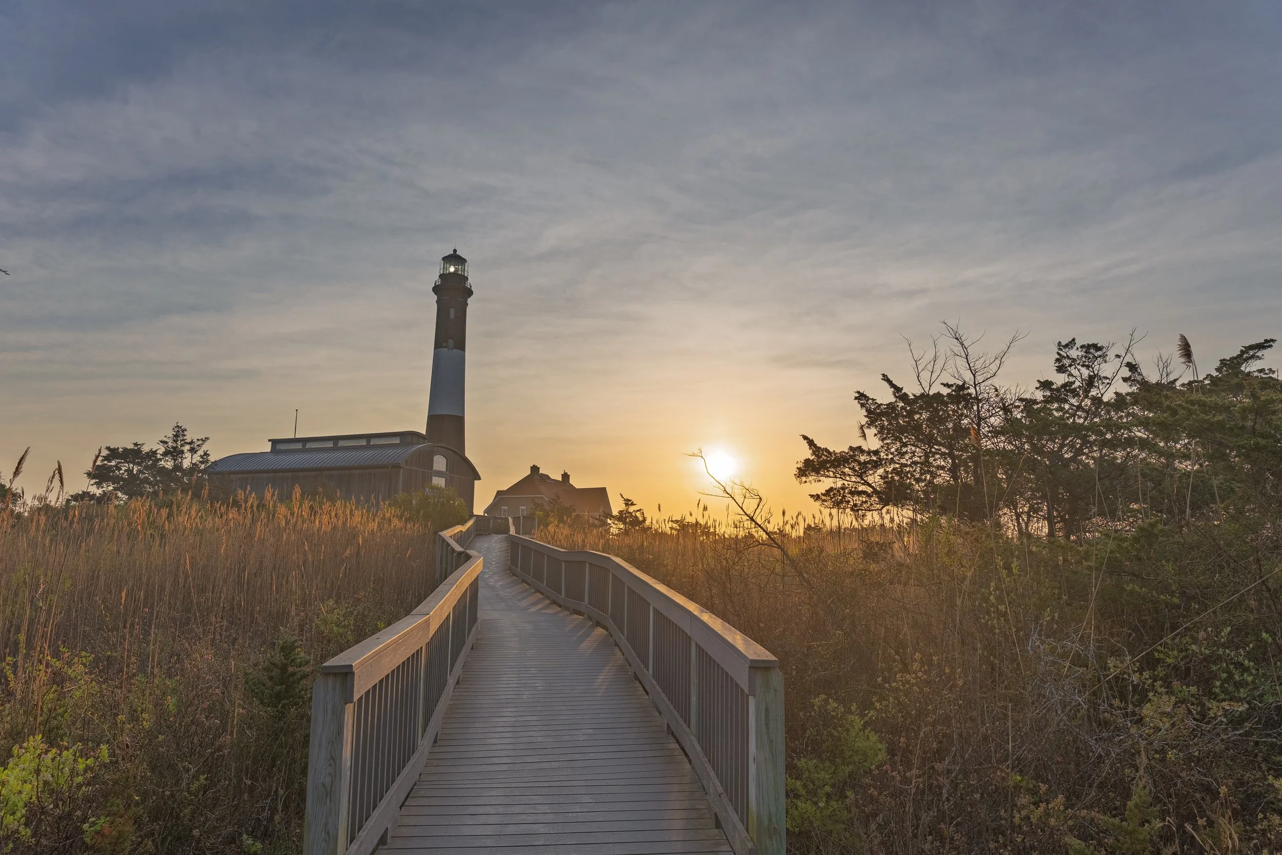 A lighthouse at sunset viewed from a wooden boardwalk leading through tall grasses and trees.