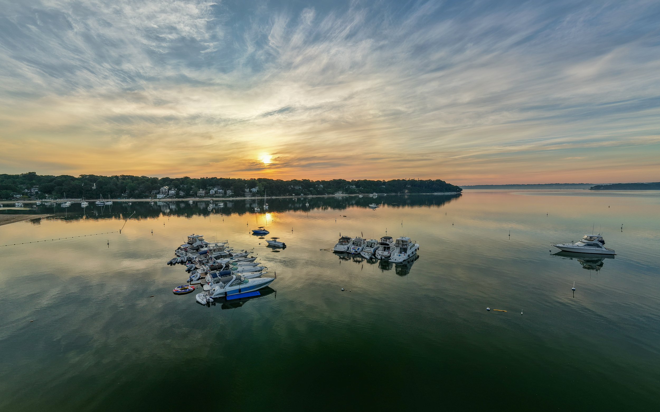 Sunset over a marina with boats docked on calm water, trees and houses in the background, and a sky filled with wispy clouds.