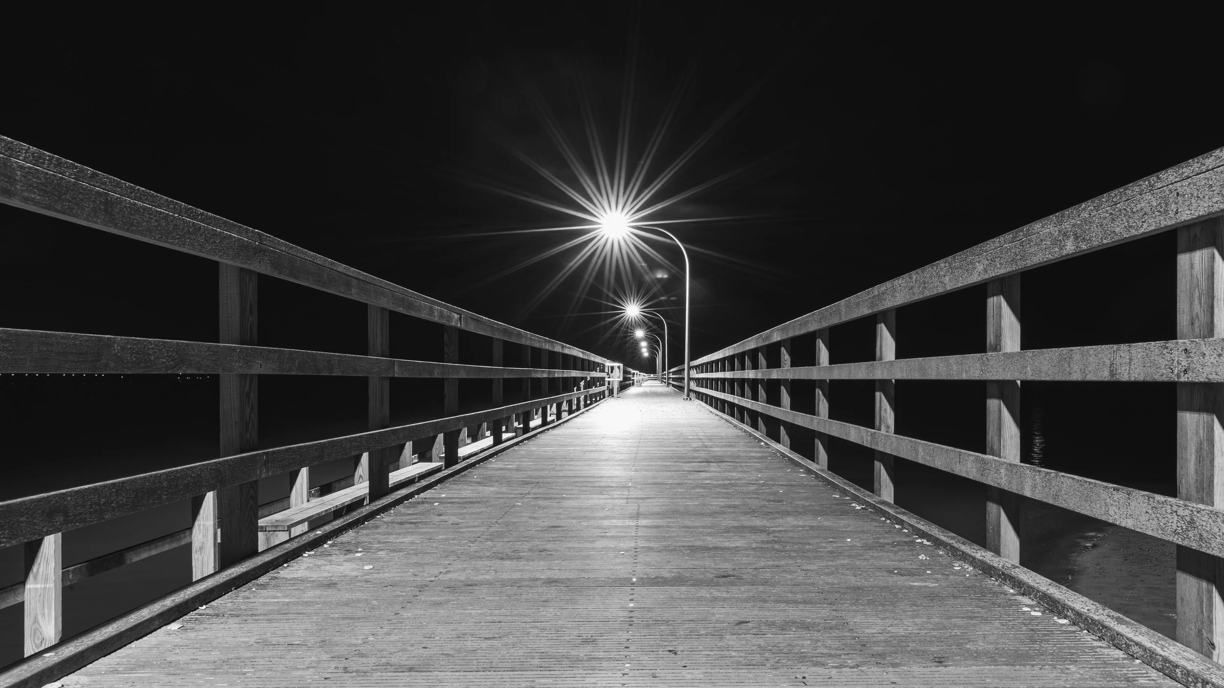 Night view of a wooden pier with railings, illuminated by streetlights, extending into darkness.