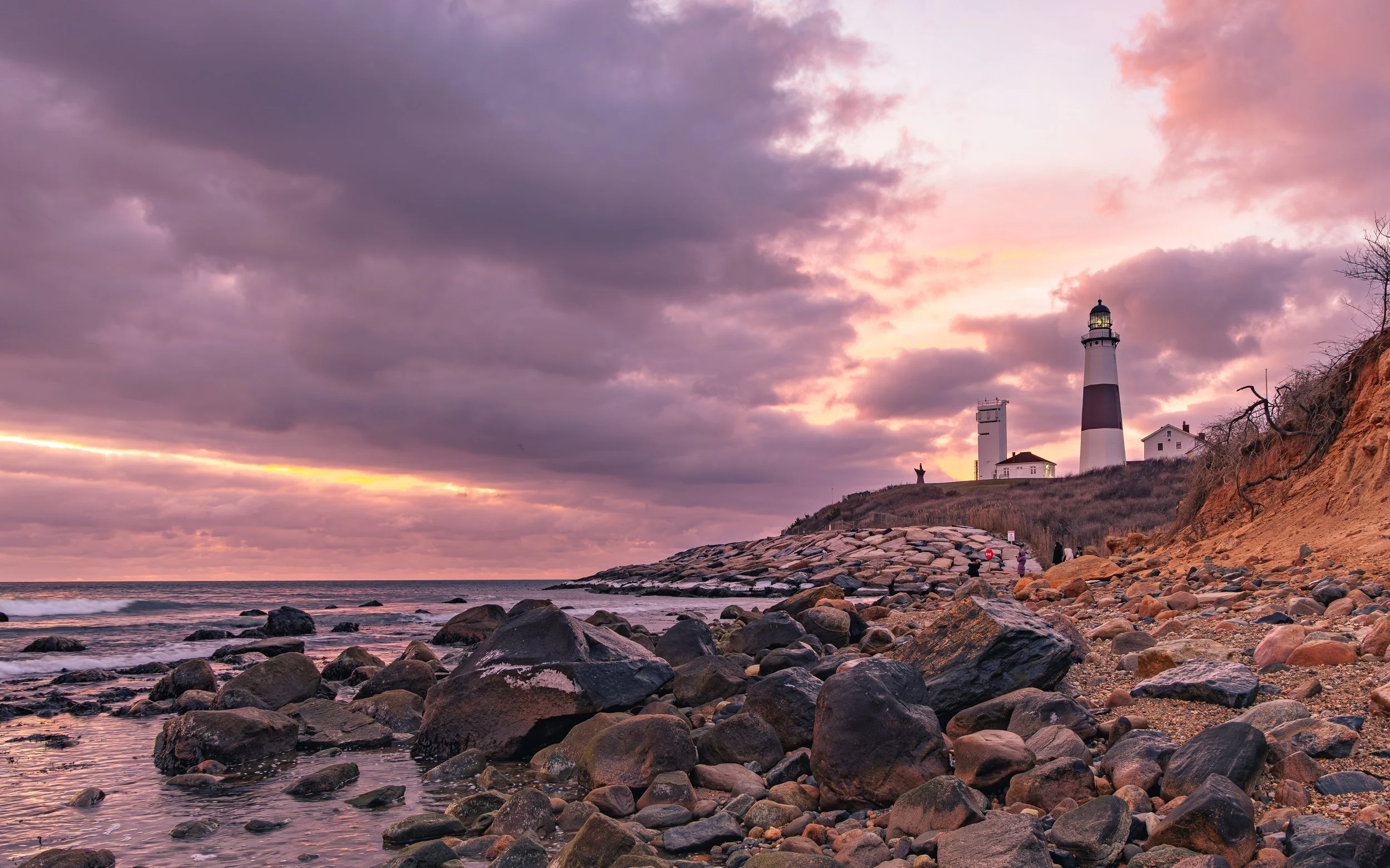 A lighthouse on a rocky coastline at sunset with a cloudy sky in shades of purple, pink, and orange, and a few people walking along the shore.