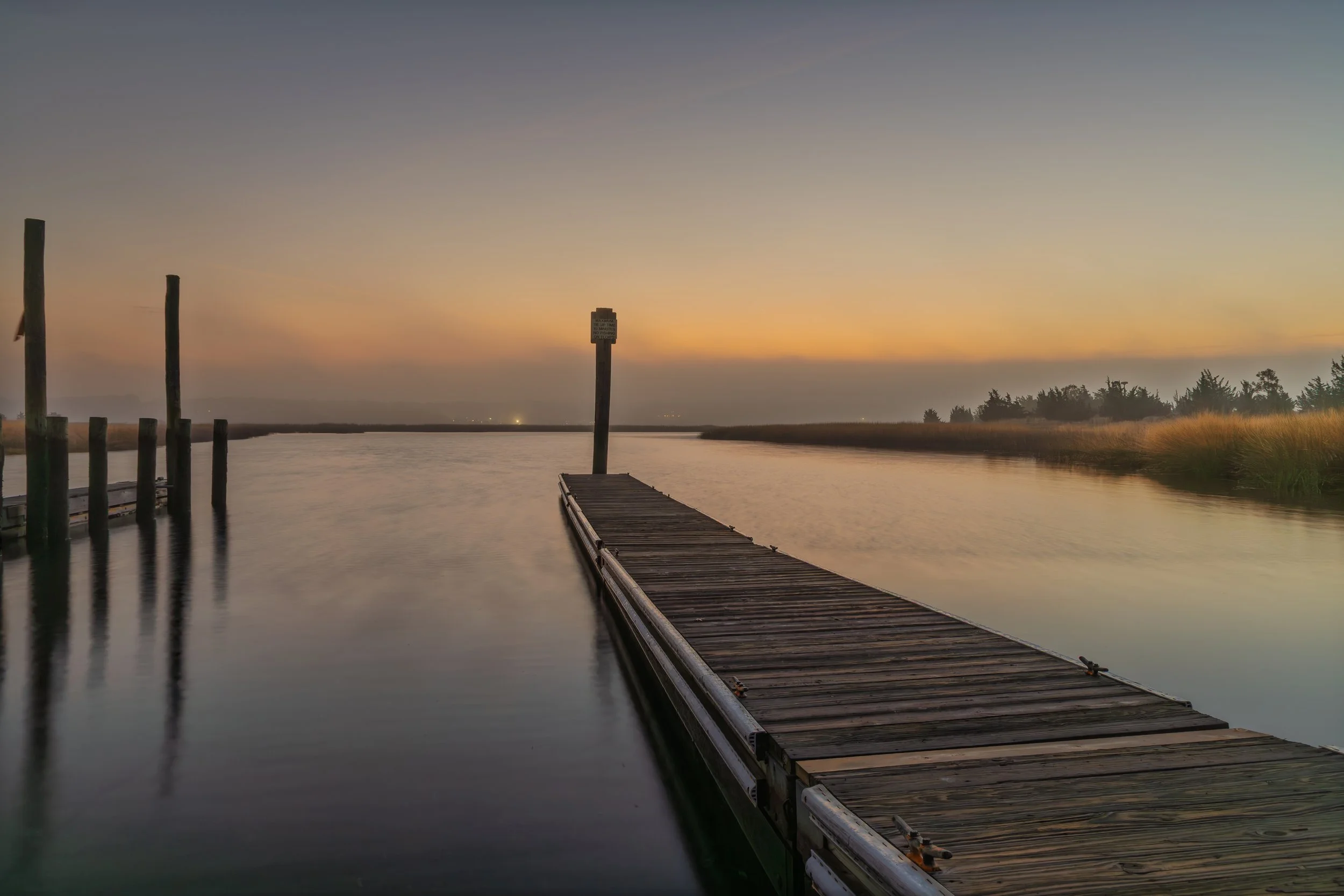 A wooden dock extends into a calm river at sunset, with a sign on one post and tall grasses on the riverbank, under a mostly clear sky.