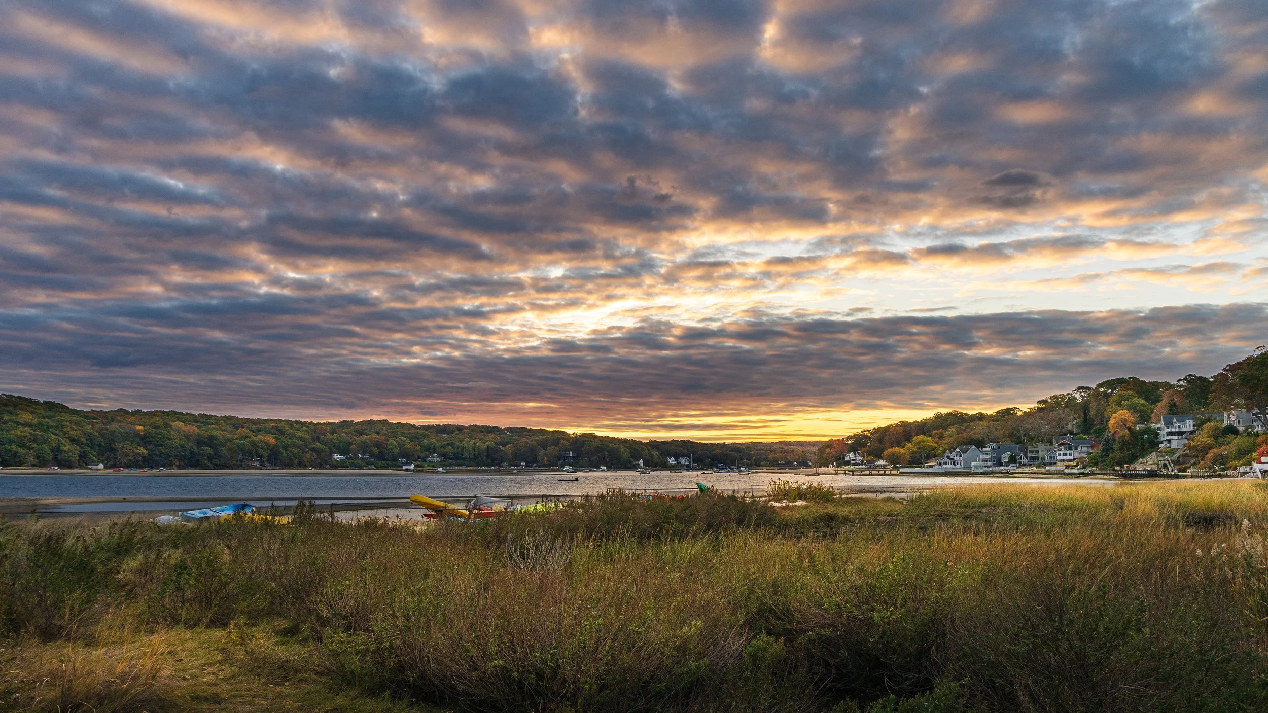 A scenic view of a river at sunset with clouds in the sky, trees on the hills, and houses along the shoreline.