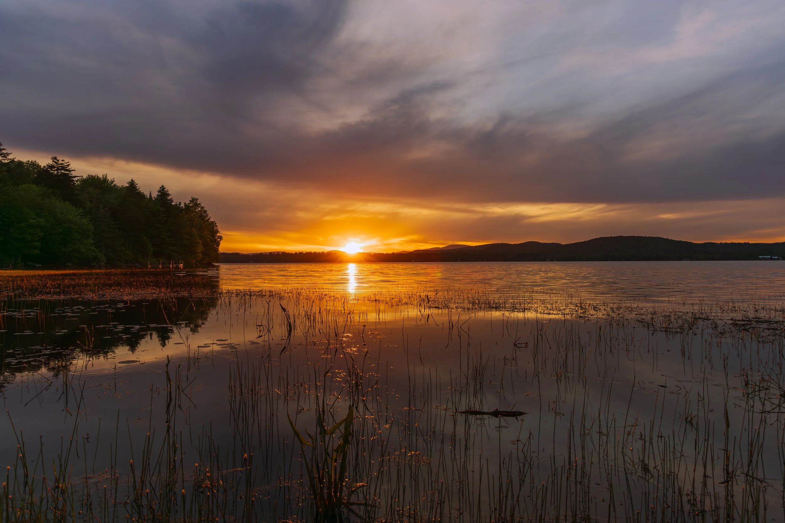 Sunset over a lake with dark clouds and reflections on the water, surrounded by trees and distant hills.