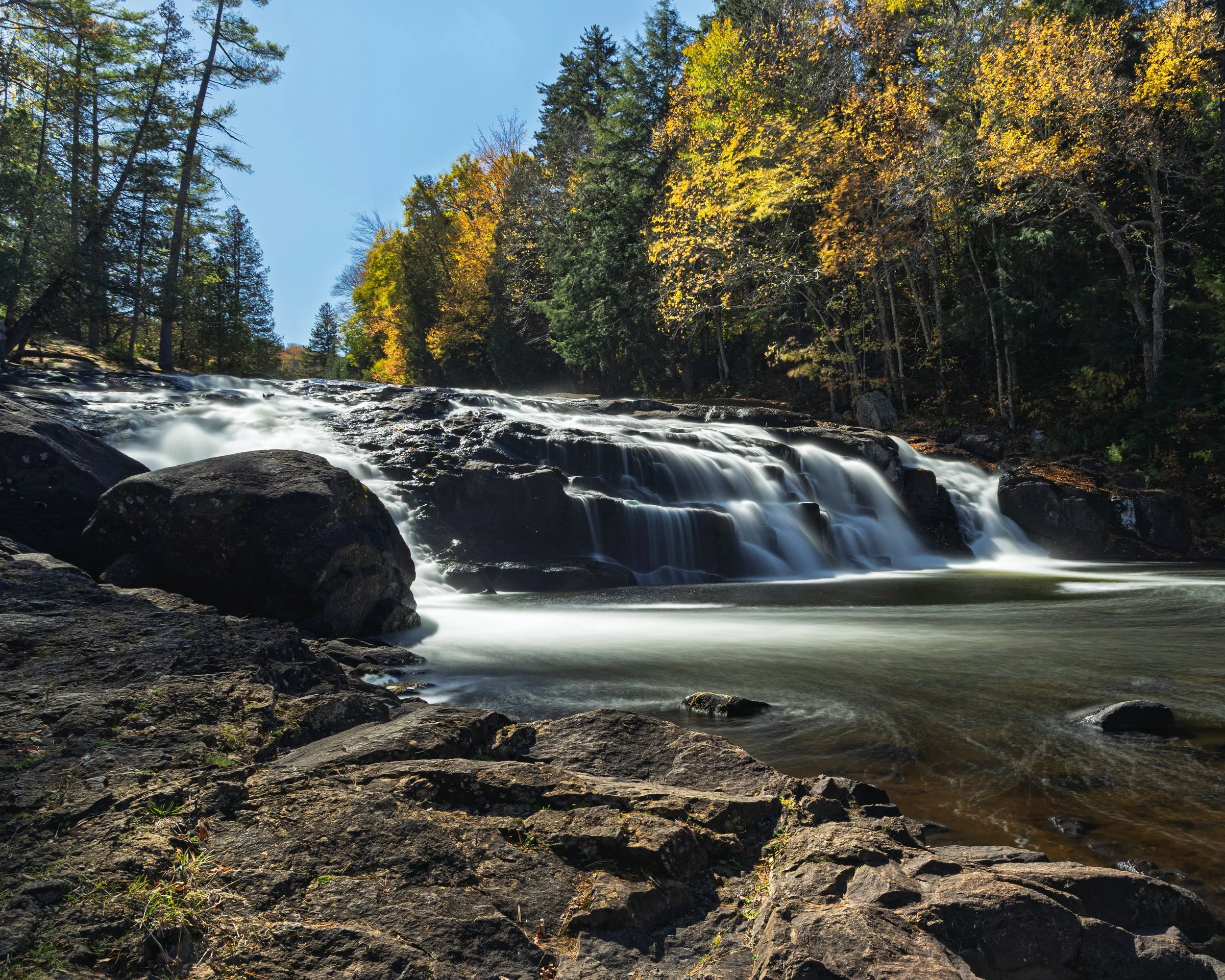 A serene waterfall flowing over rocks in a forest with trees showing fall foliage, blue sky, and sunlight filtering through the leaves.
